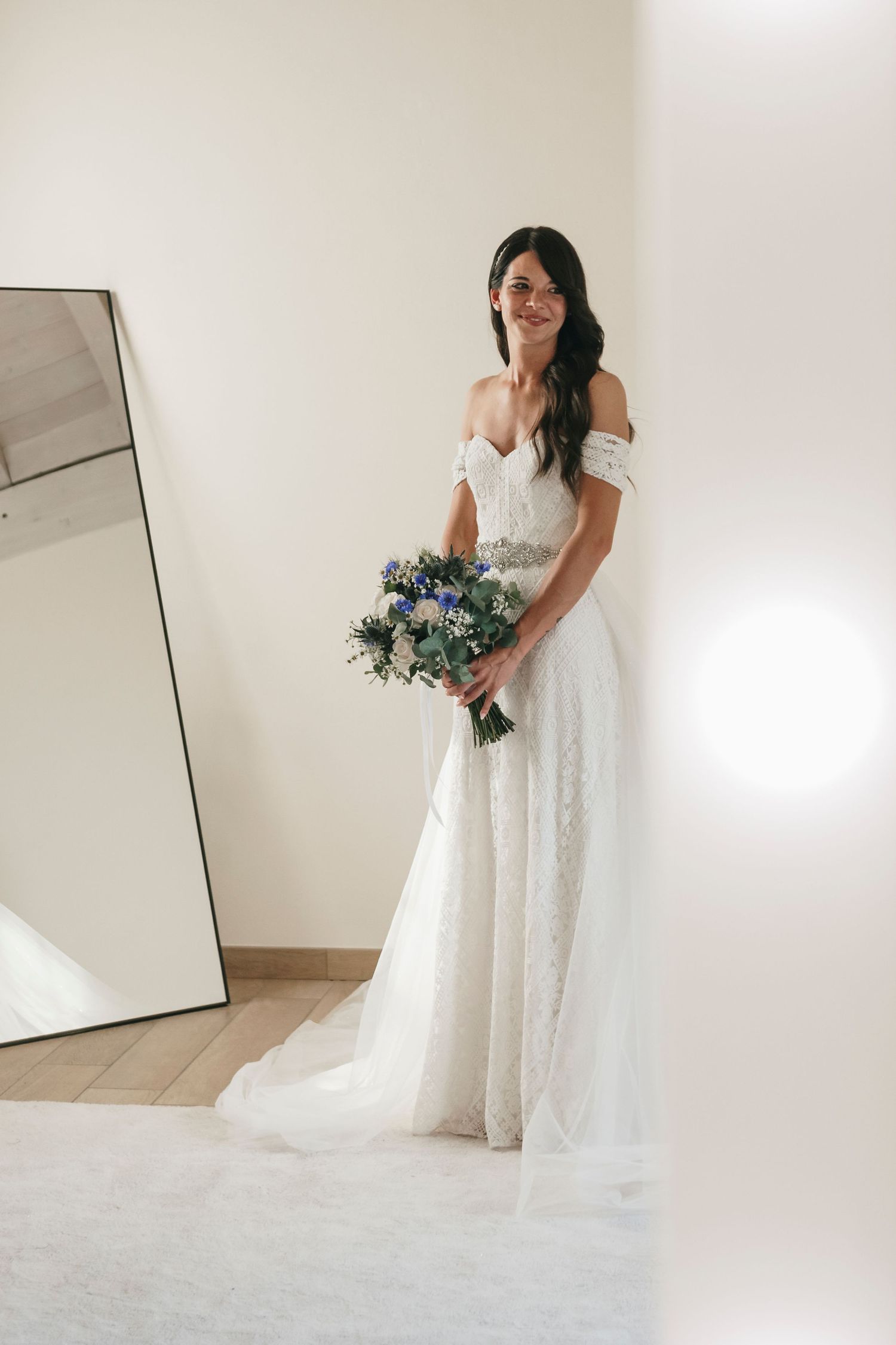 Smiling bride in off-shoulder lace wedding gown holds blue bouquet beside floor mirror in bright room.