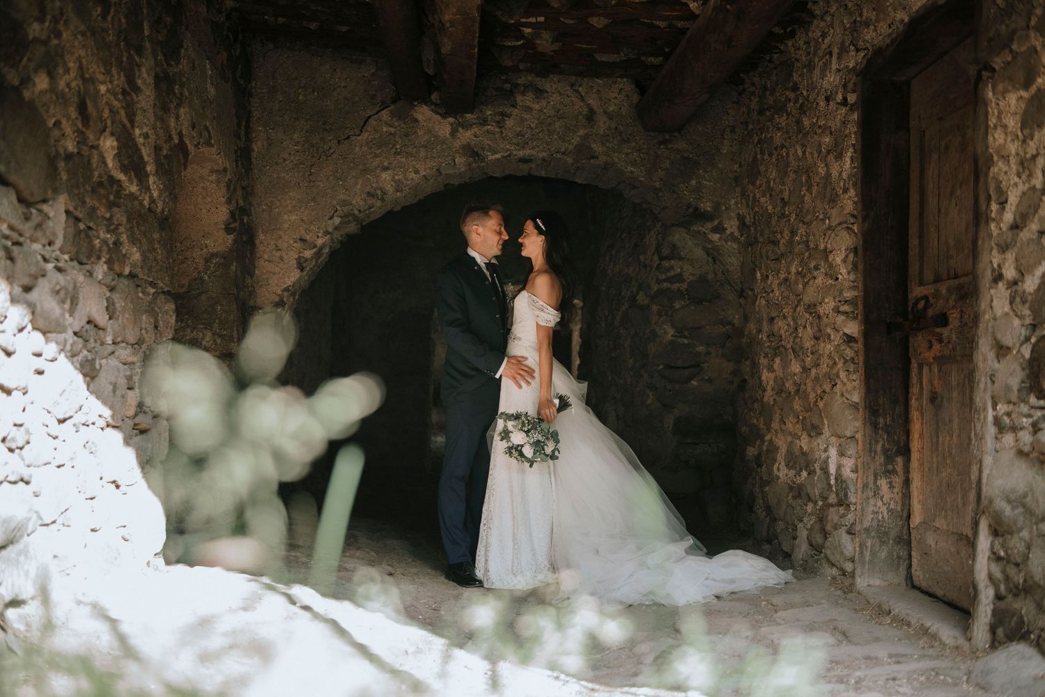 Bride and groom share a romantic moment in a rustic stone archway, bride holding a bouquet in an elegant white gown.
