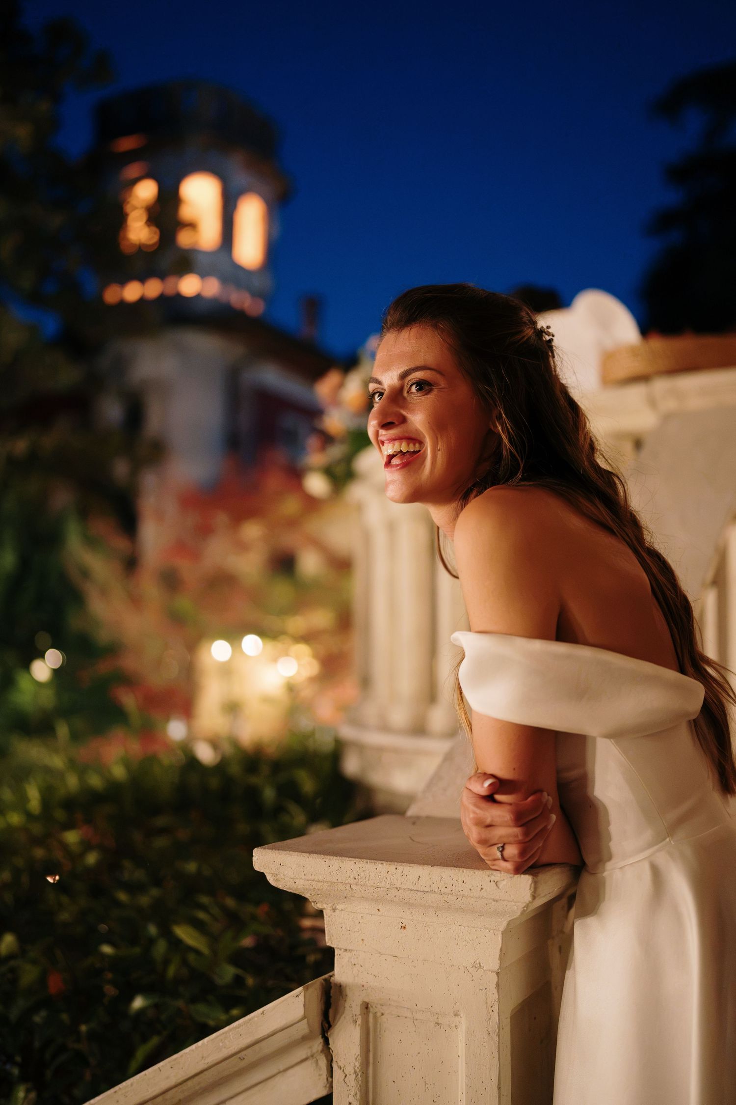 Bride in off-shoulder white gown smiling on balcony at night with illuminated historic building backdrop.