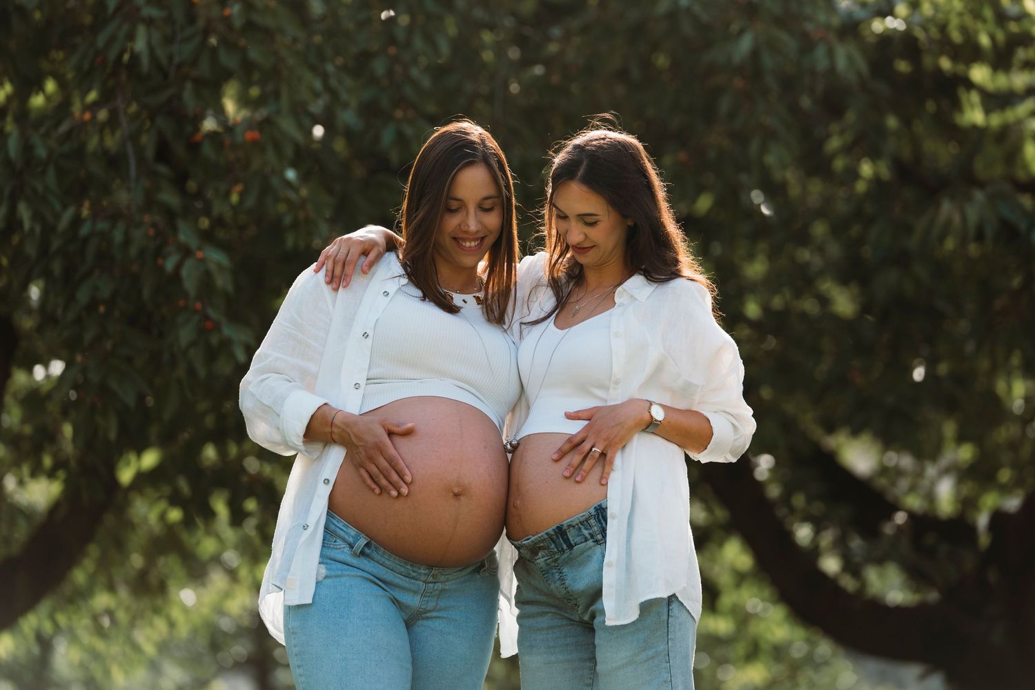 Two pregnant women in white shirts and jeans smiling and cradling their baby bumps outdoors.