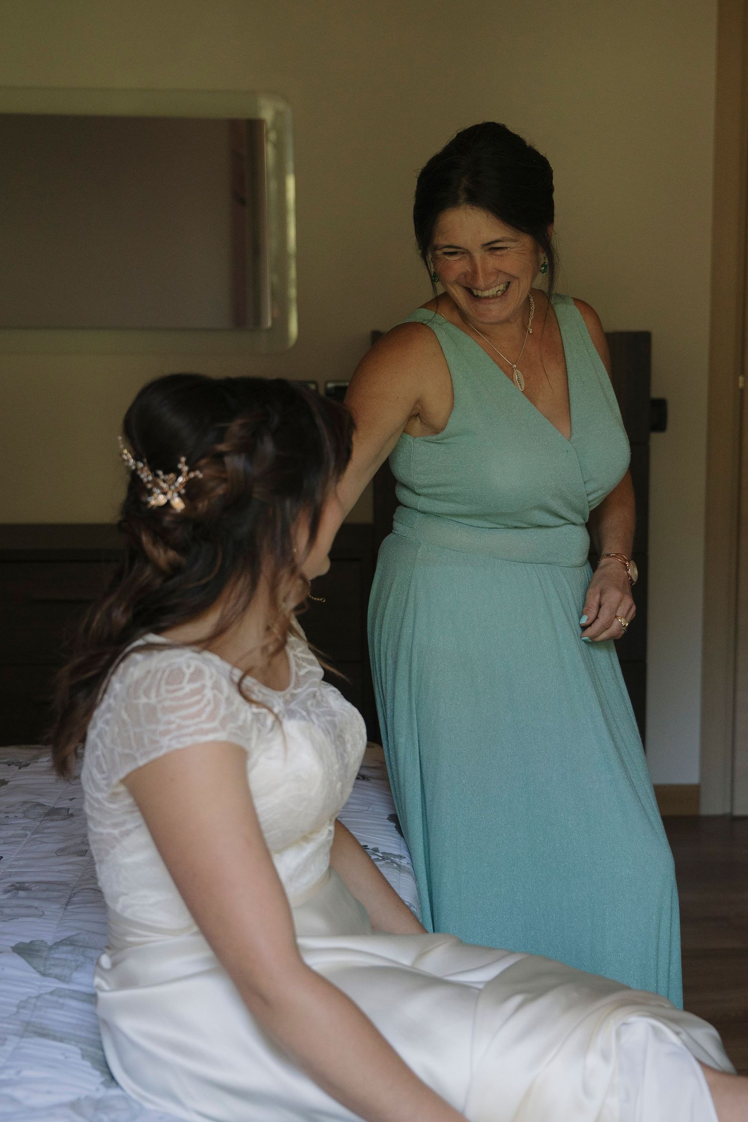 Mother smiling joyfully while helping bride in white lace wedding dress prepare for the ceremony.