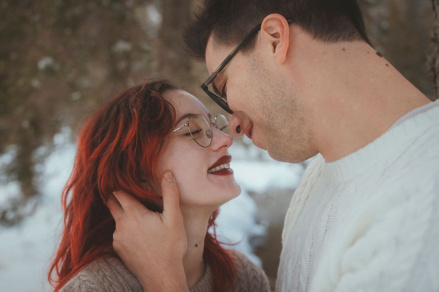 Couple almost kissing in a snowy winter forest, woman with vibrant red hair and glasses.