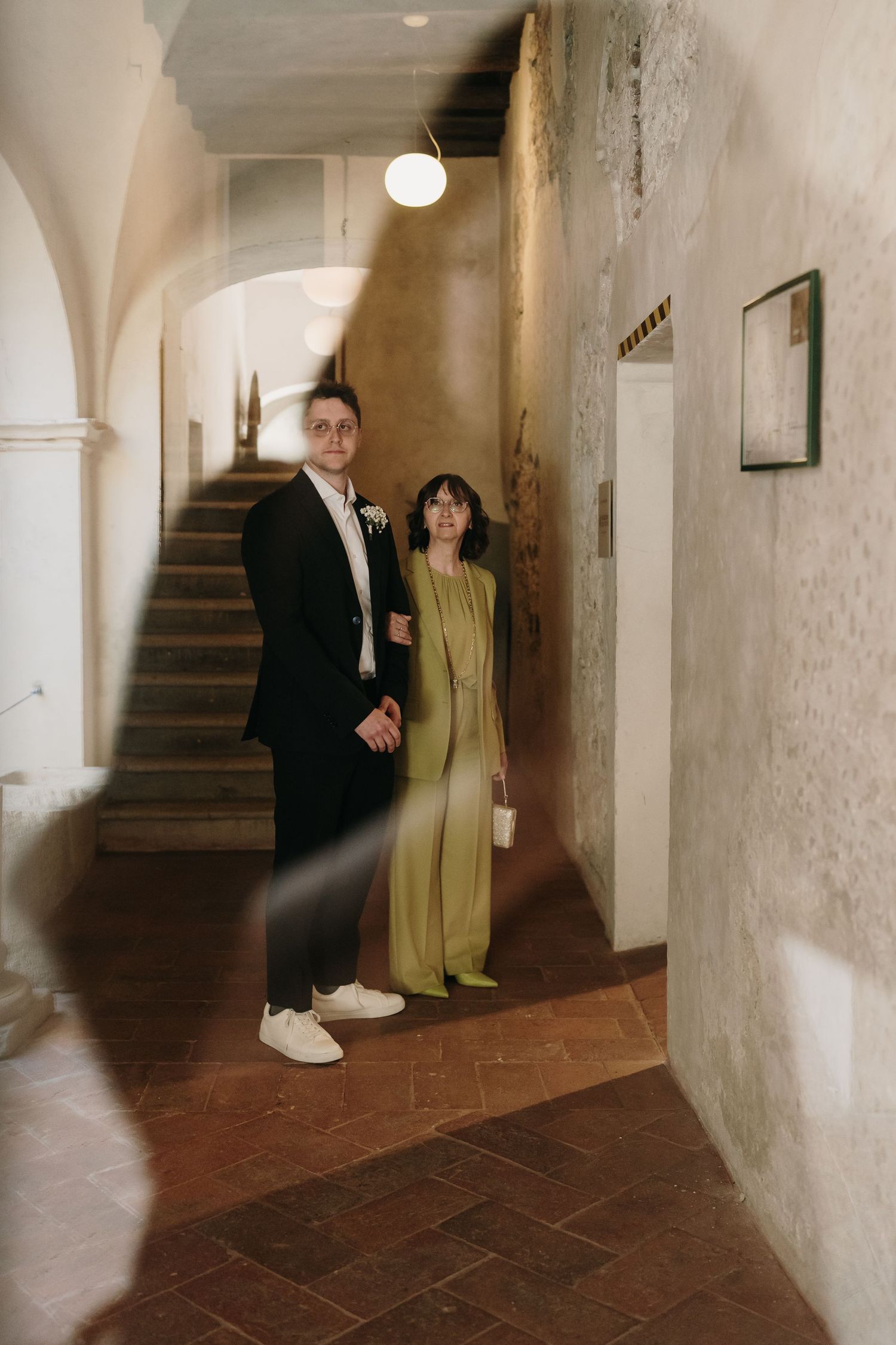 Couple in formal attire poses in a rustic arched corridor with terracotta floors and stone walls.