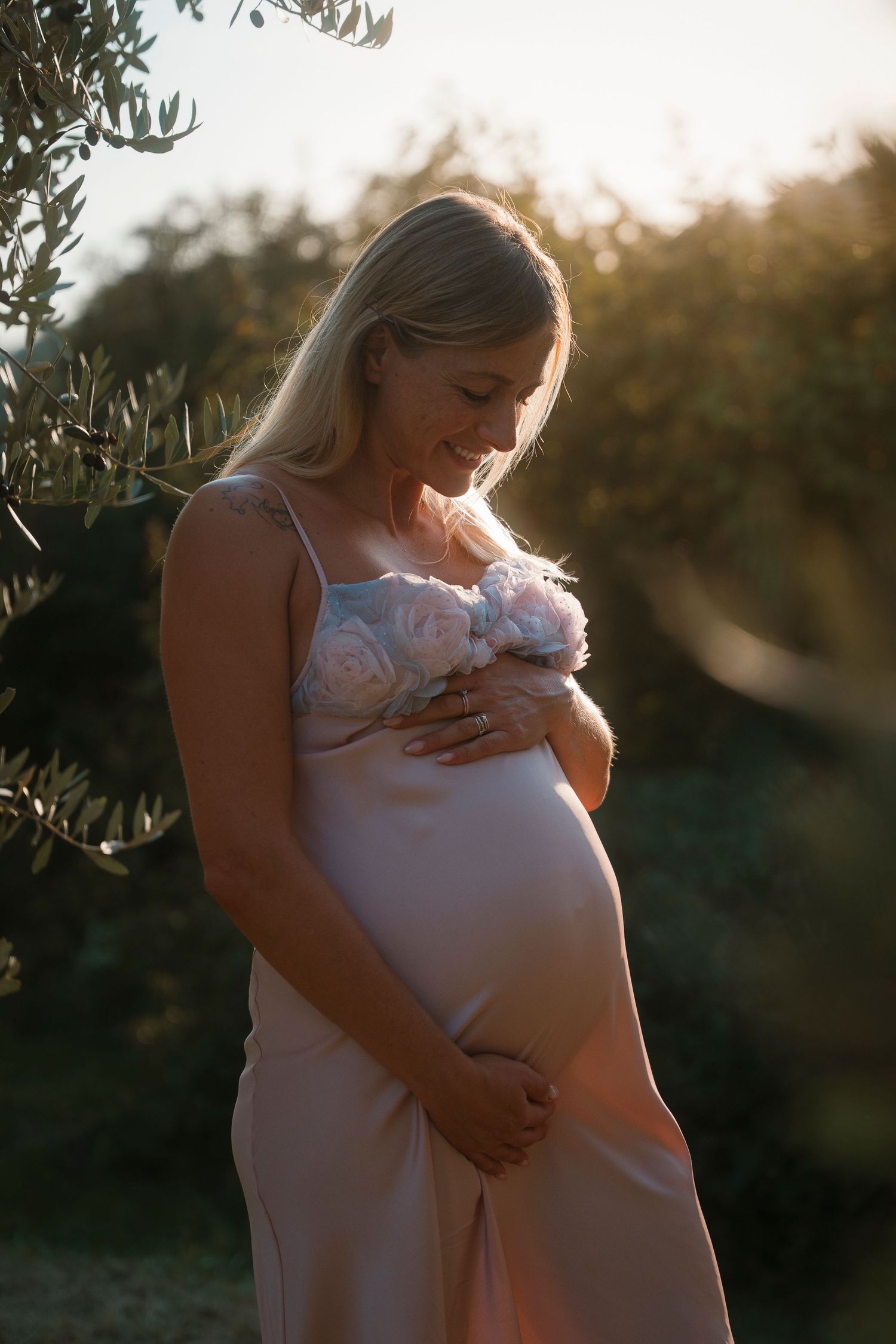 Pregnant blonde woman in pink satin dress cradling her bump among olive trees at sunset.