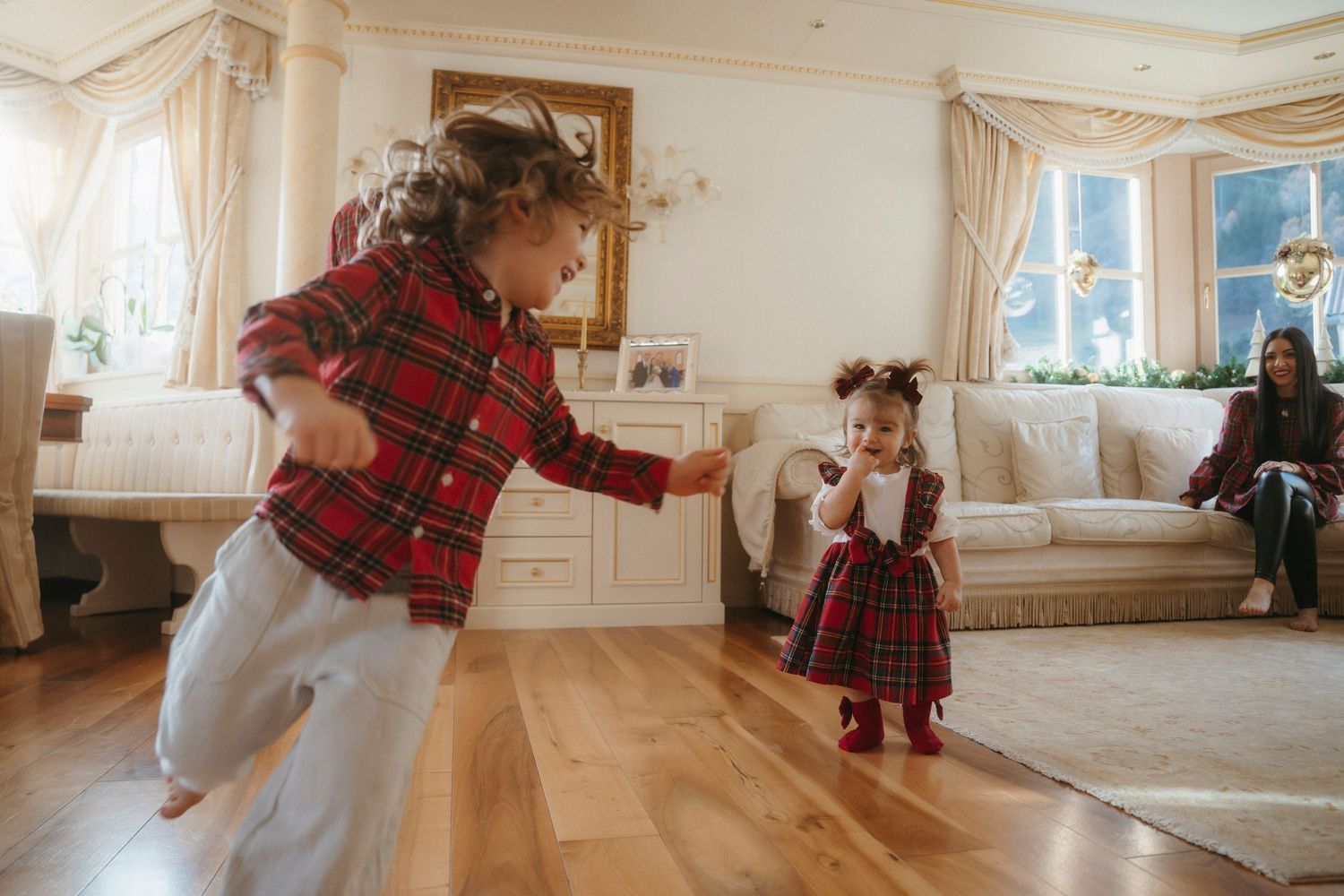 Children in red plaid outfits dancing and playing on hardwood floor in festive living room.