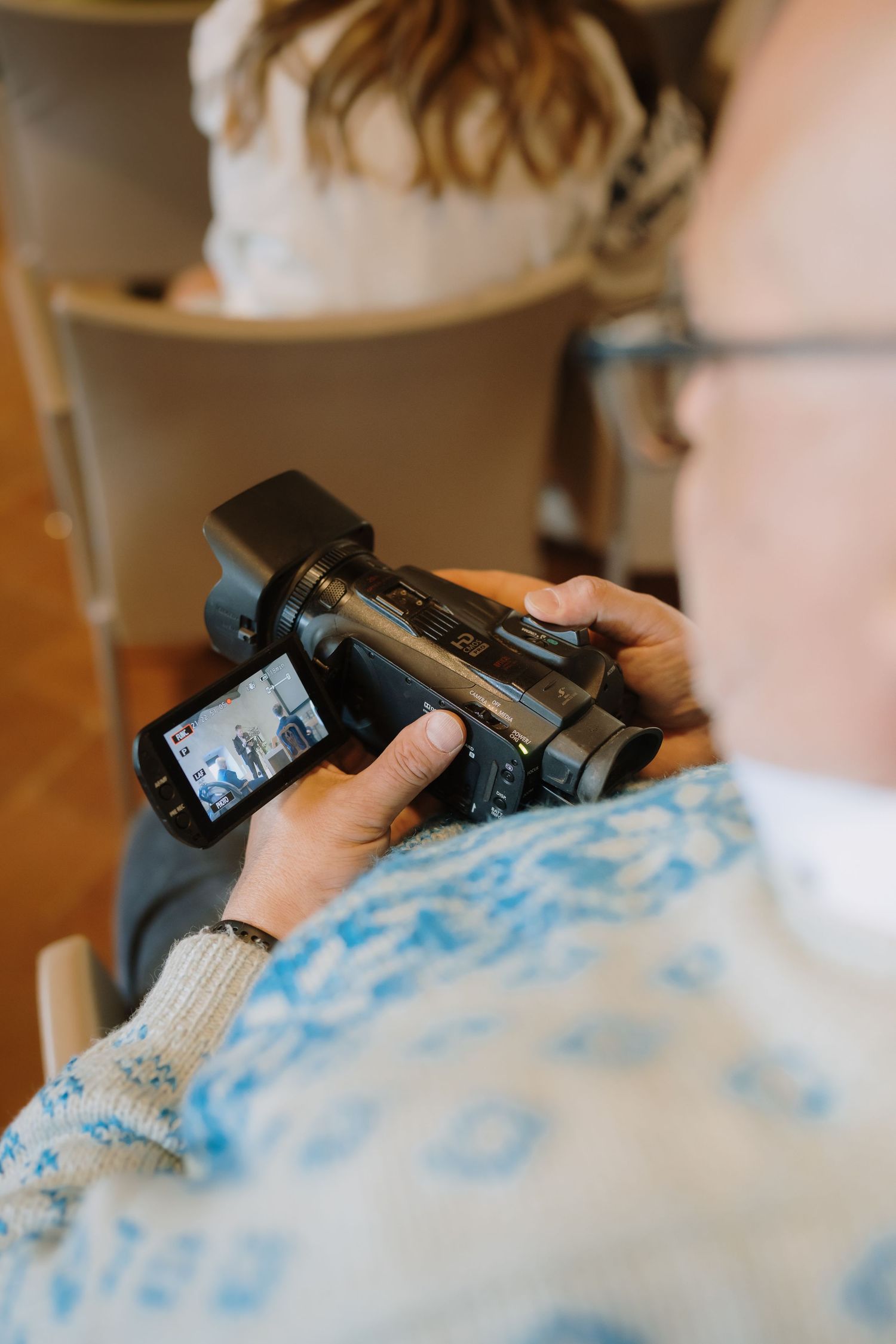 Person in blue sweater holding a video camera and reviewing footage on the flip screen at an indoor event.