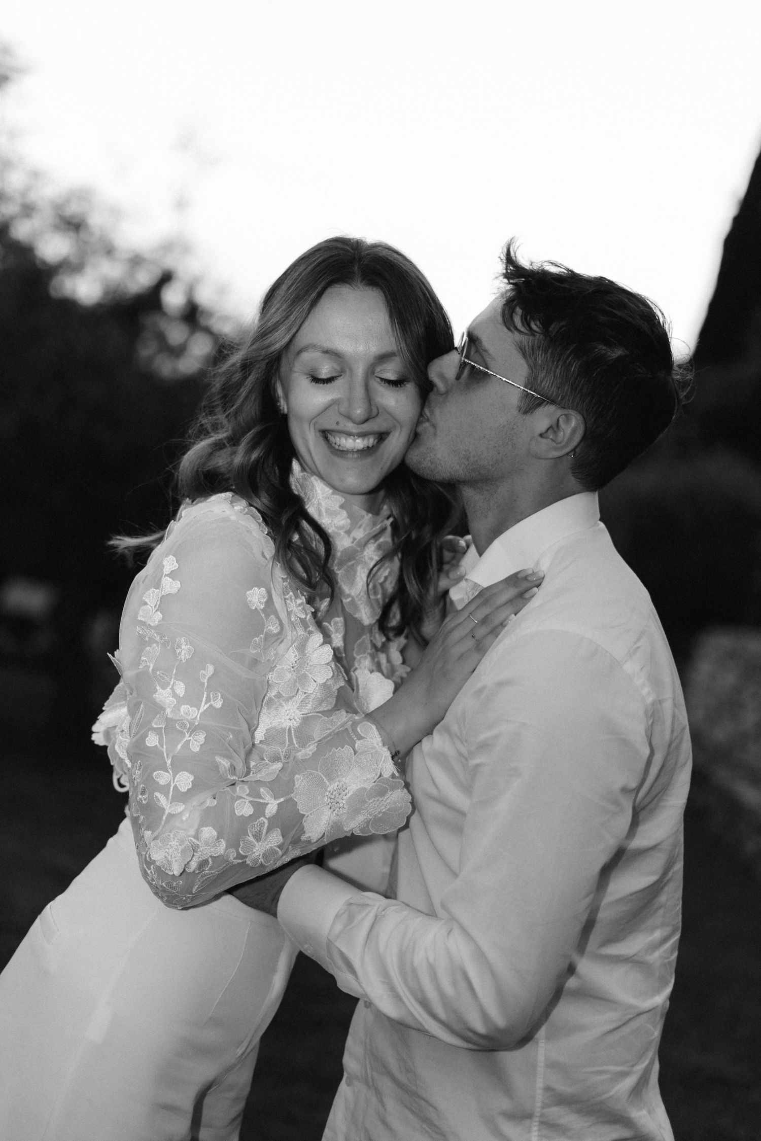 Groom kisses smiling bride on cheek in romantic black and white outdoor wedding portrait.