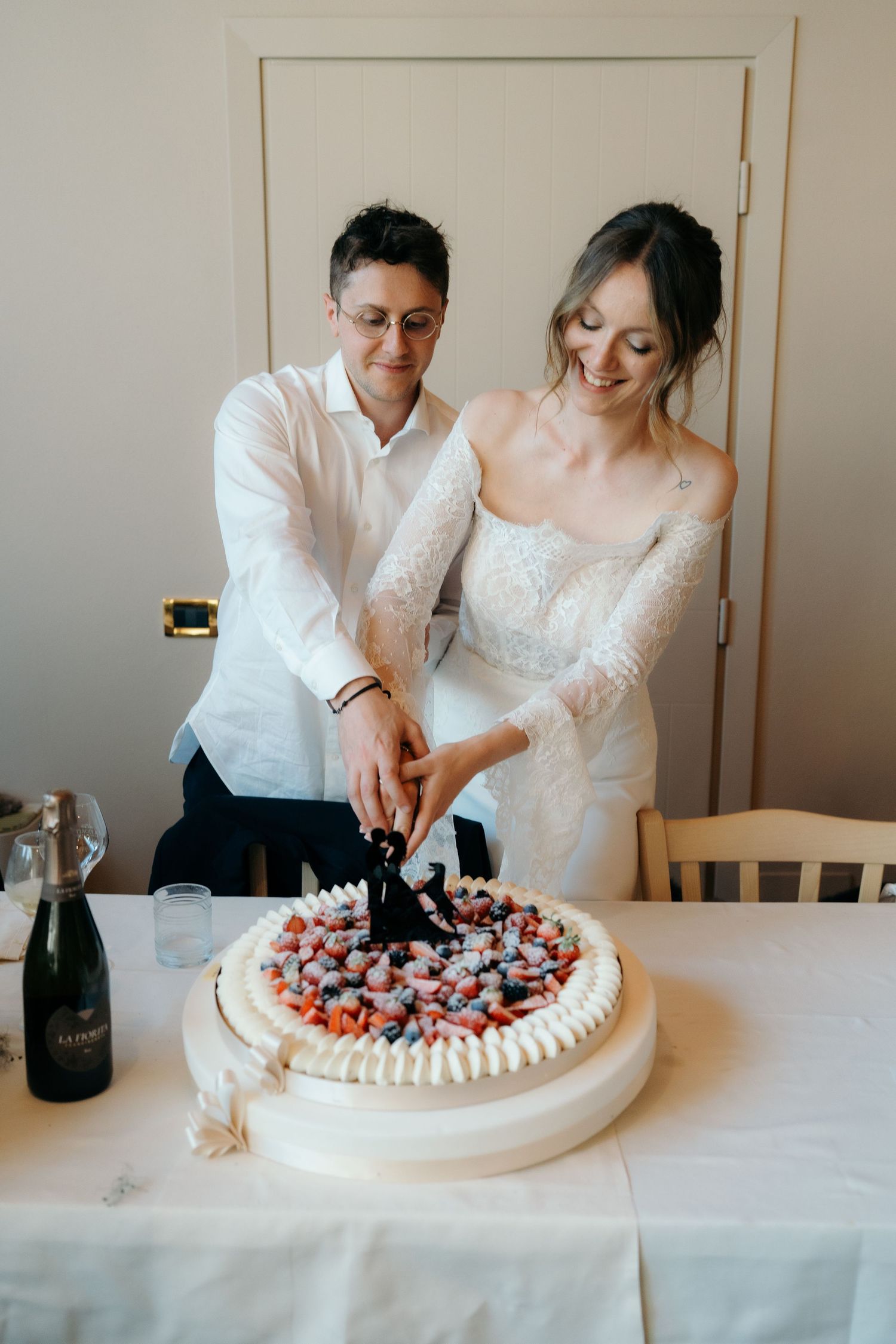 Sposi sorridenti in abiti bianchi tagliano insieme una torta nuziale rotonda guarnita con frutti di bosco e fragole fresche.