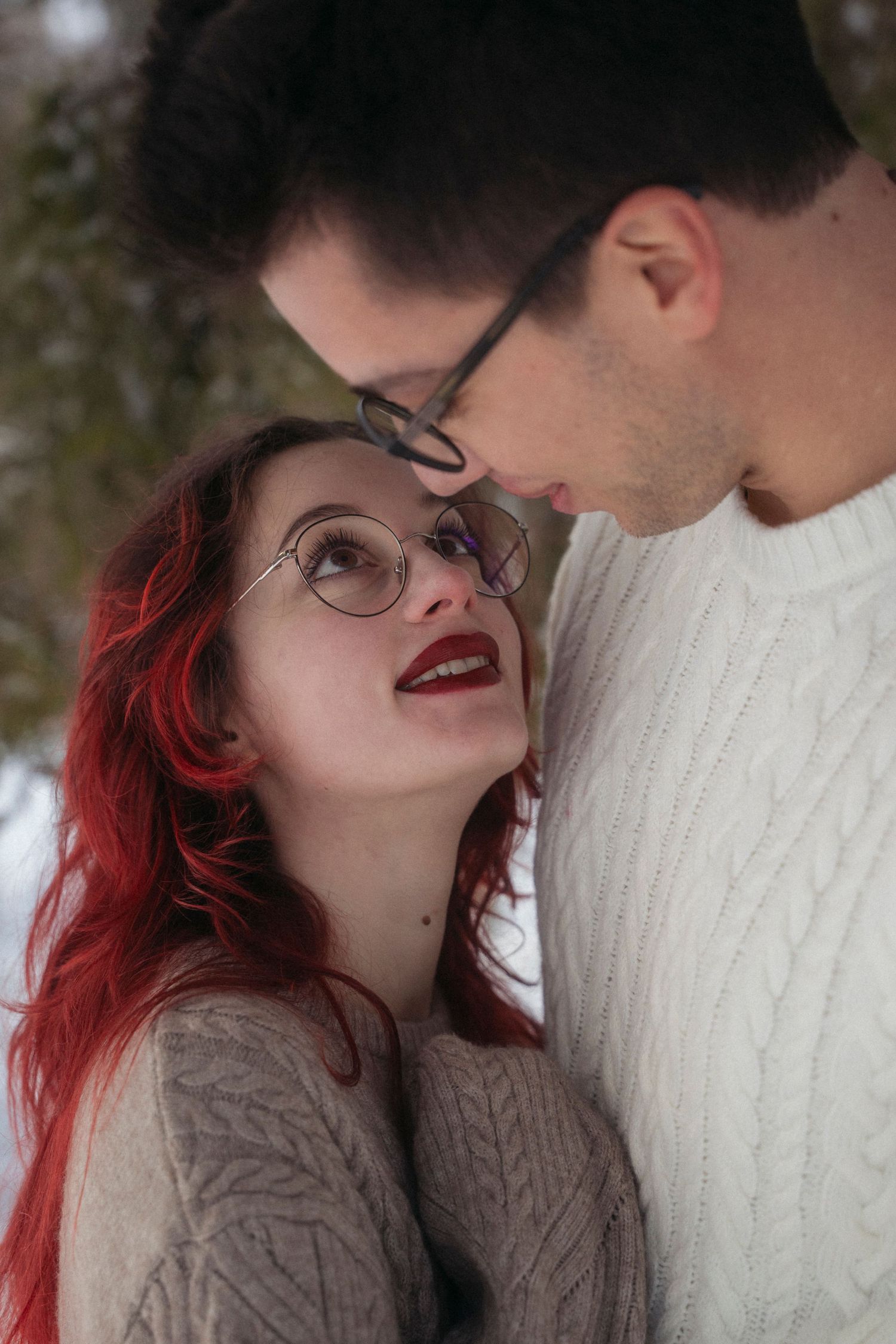 A couple in matching knit sweaters share an intimate moment in a snowy winter forest, the woman with red hair and glasses smiling up.