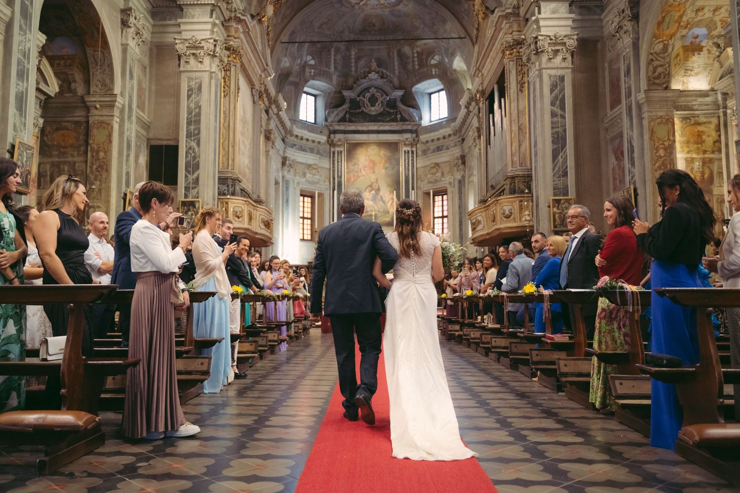 Bride walking down the red carpet aisle of an ornate church with guests applauding.