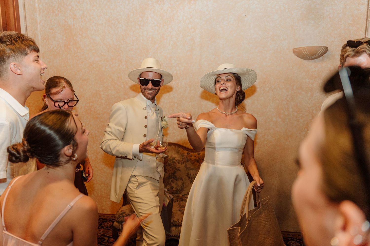 Bride and groom in white hats laughing and dancing with guests at a wedding reception.