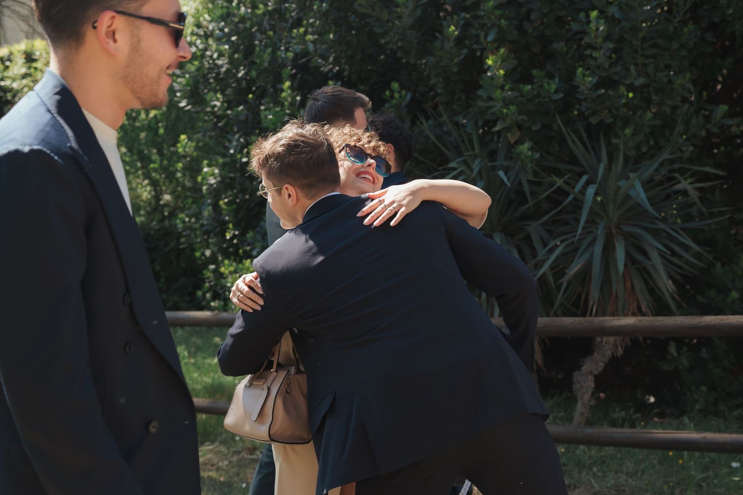 Group of young men in formal attire sharing a warm embrace outdoors near lush green bushes.