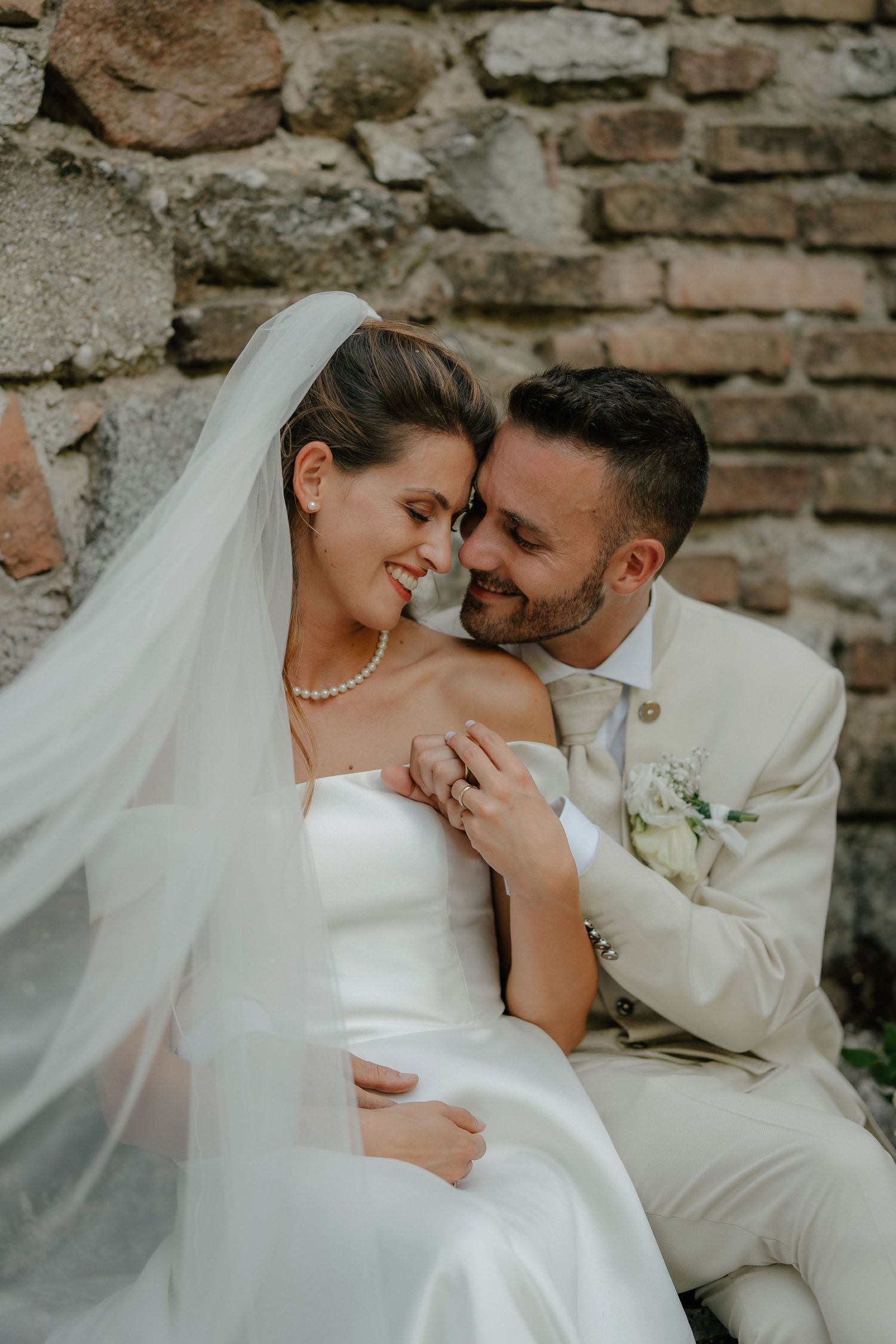 A bride and groom in white attire share a romantic moment seated against a rustic stone wall on their wedding day.
