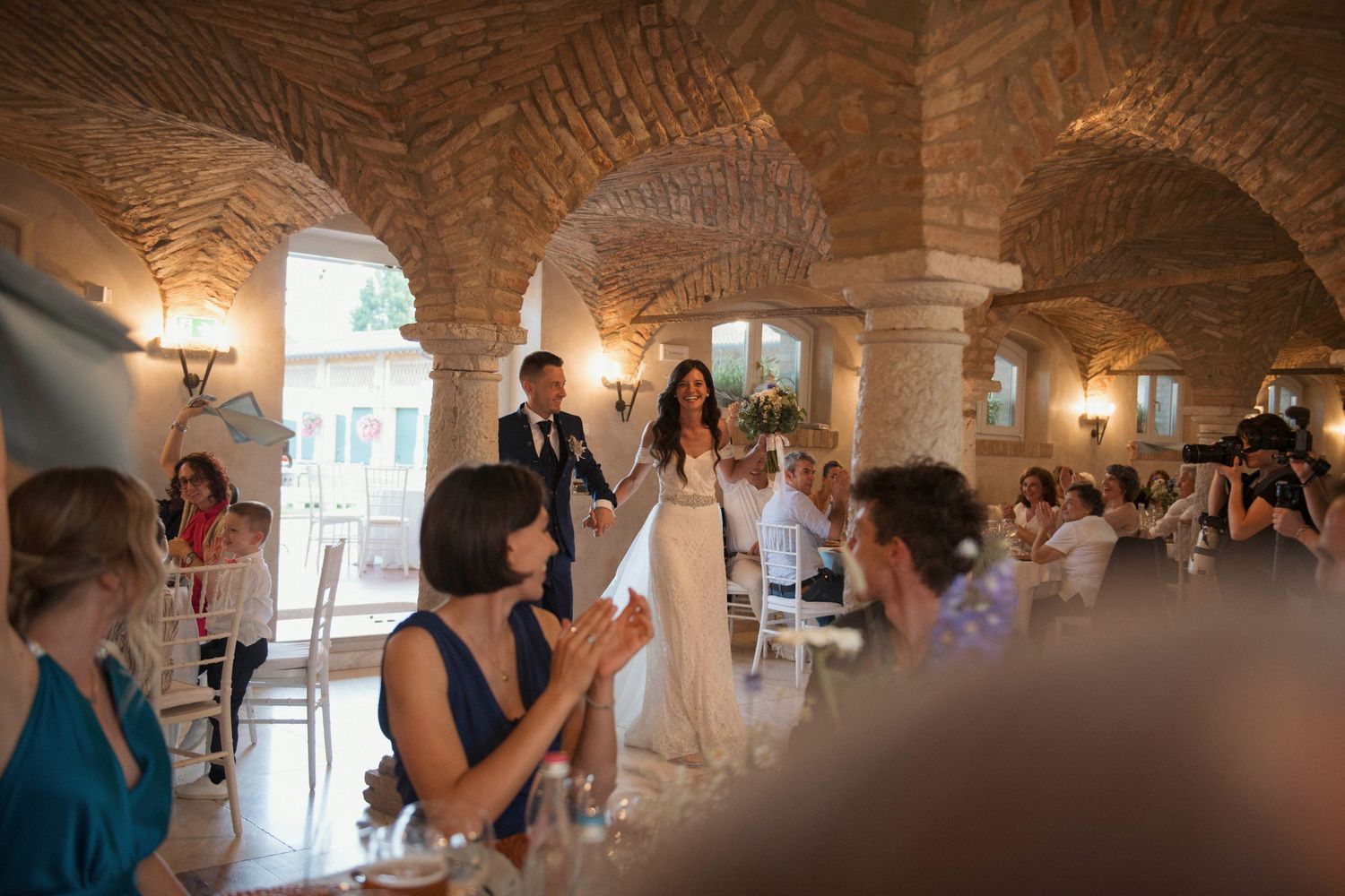Newlyweds enter rustic stone-vaulted reception hall as guests applaud and celebrate.