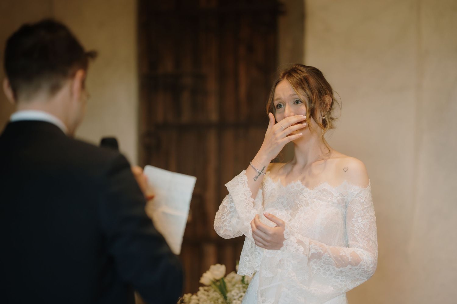 Una sposa commossa, in abito di pizzo, si copre la bocca mentre ascolta le promesse dello sposo durante la cerimonia.
