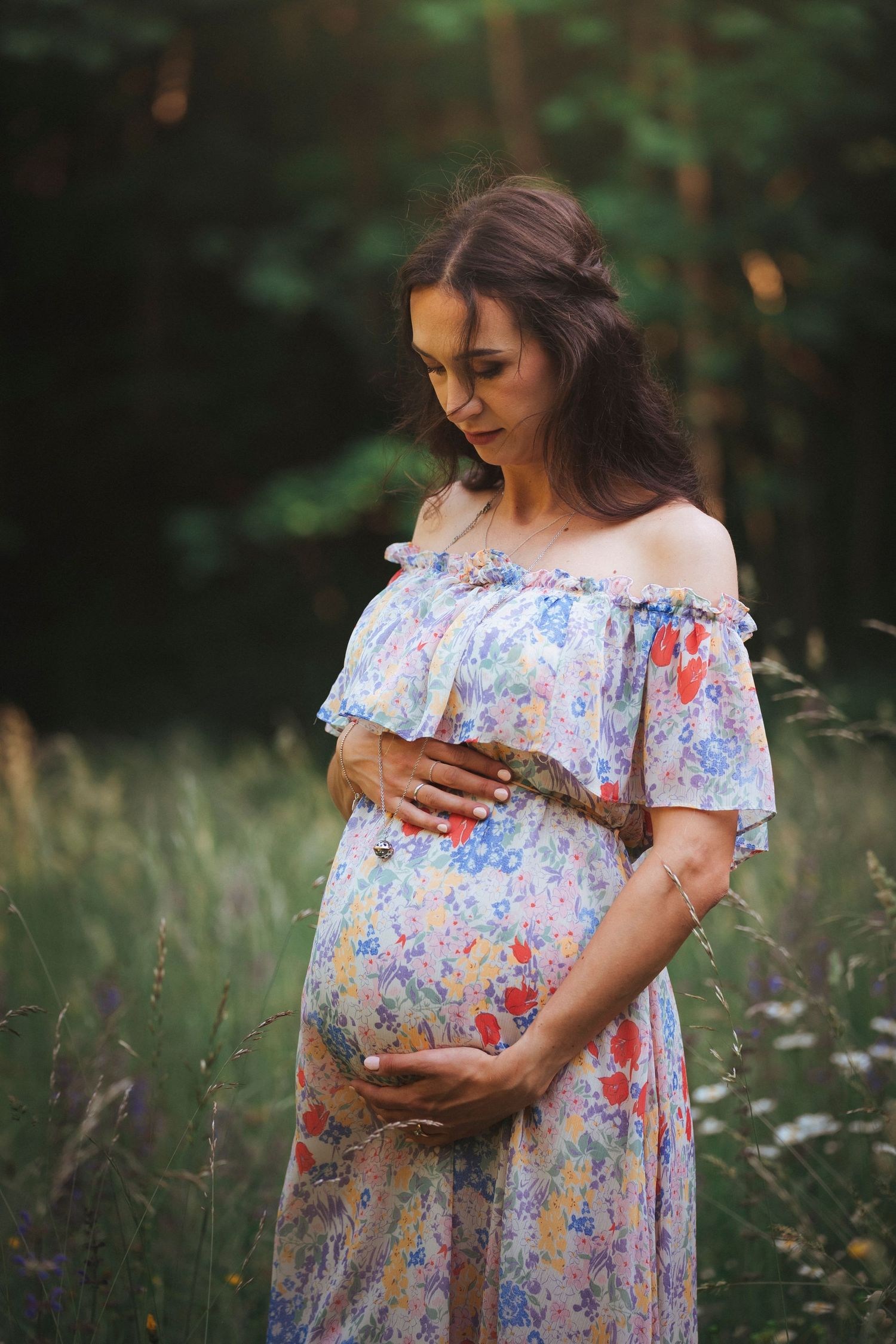 Pregnant woman in a floral dress gently cradling her baby bump in a serene outdoor meadow.