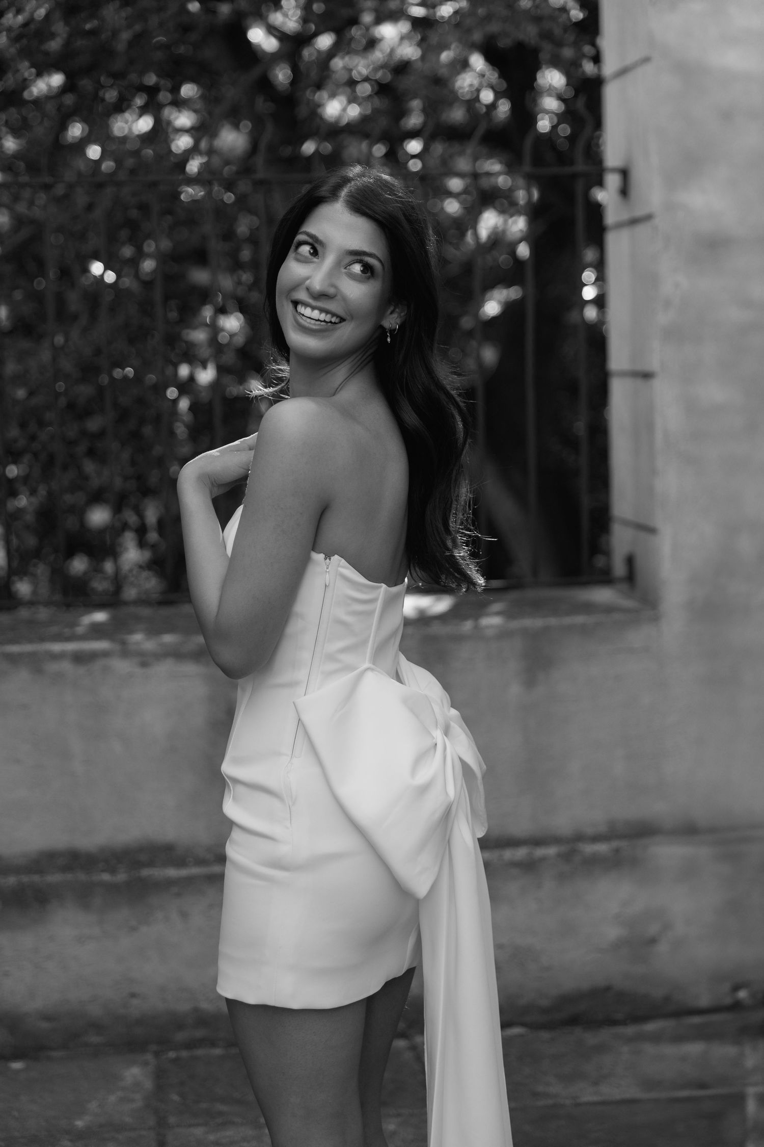 A black and white portrait of a bride in a white mini dress with a dramatic bow detail.