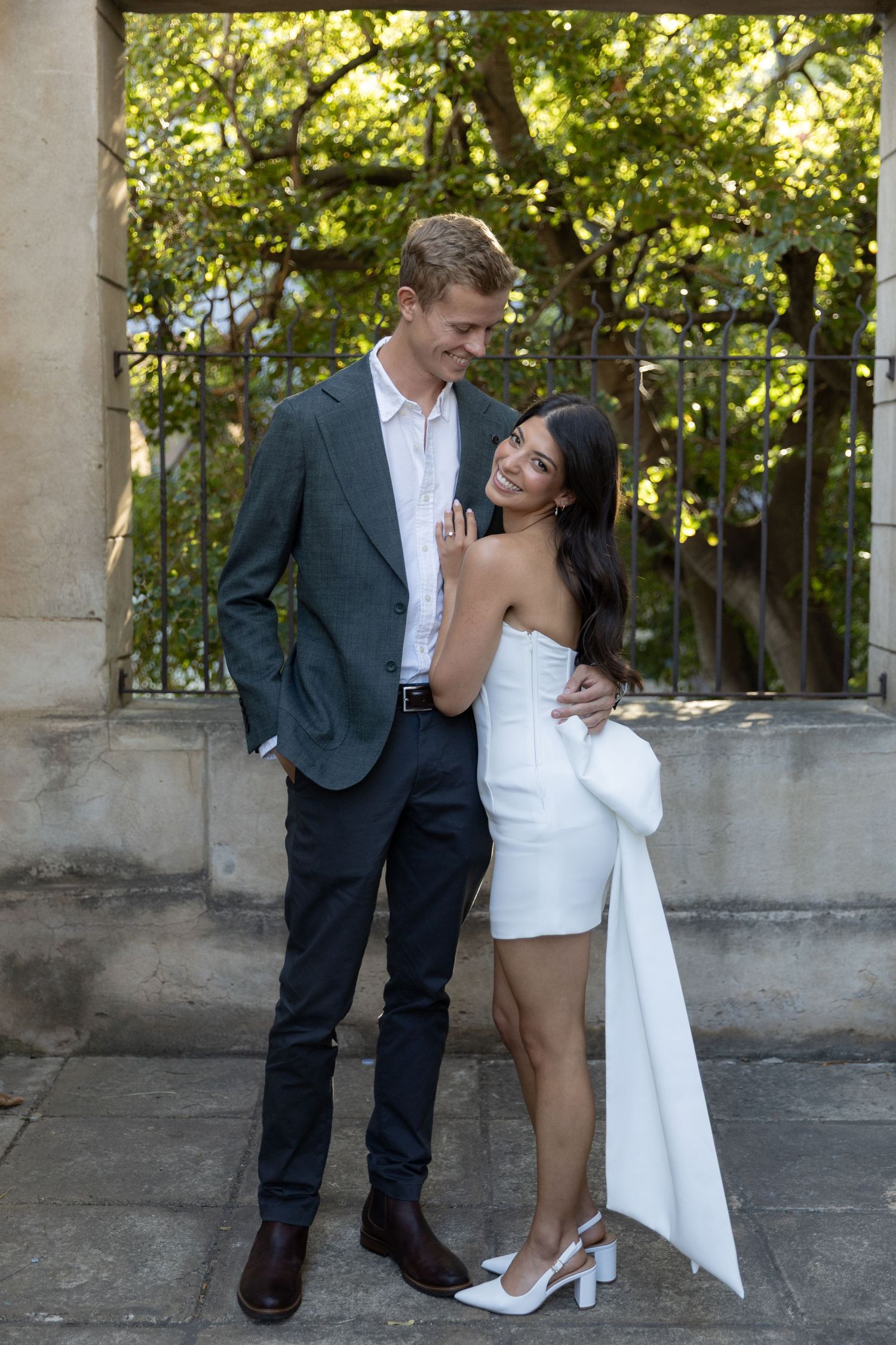 A couple laughs together on a balcony, she in a white dress with train and he in a suit.