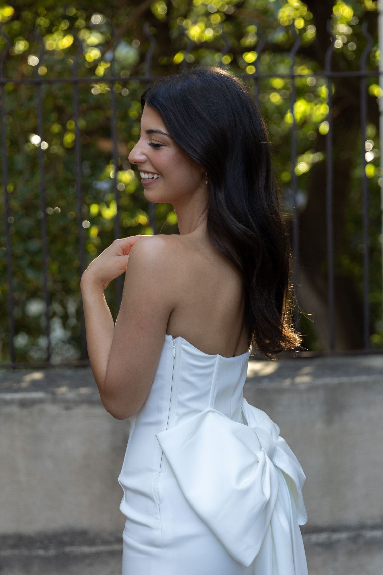 Woman in white strapless dress poses with flowing dark hair against garden backdrop.