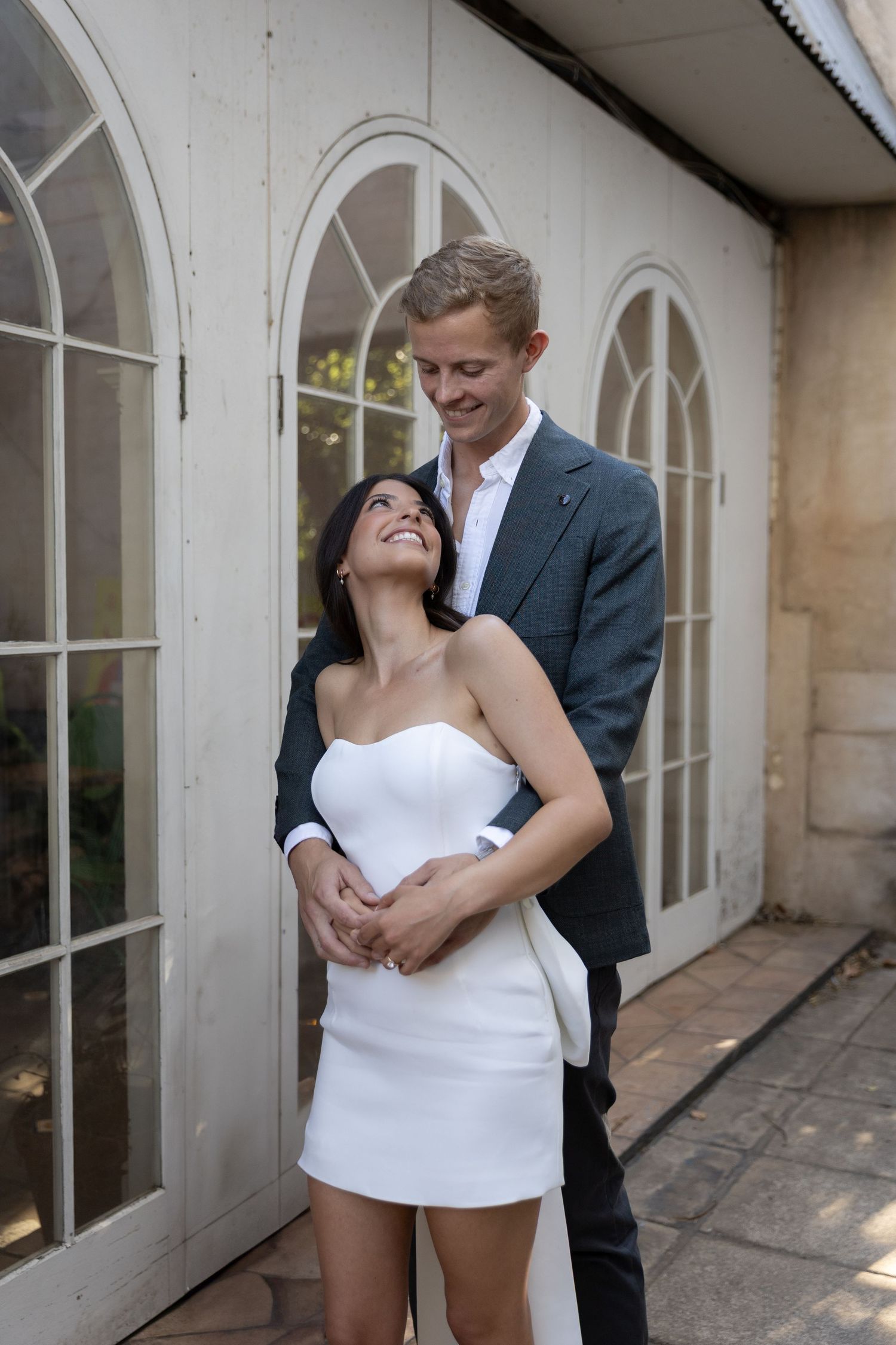 Couple sharing an intimate moment in front of arched glass doors, with the woman wearing a white mini dress.