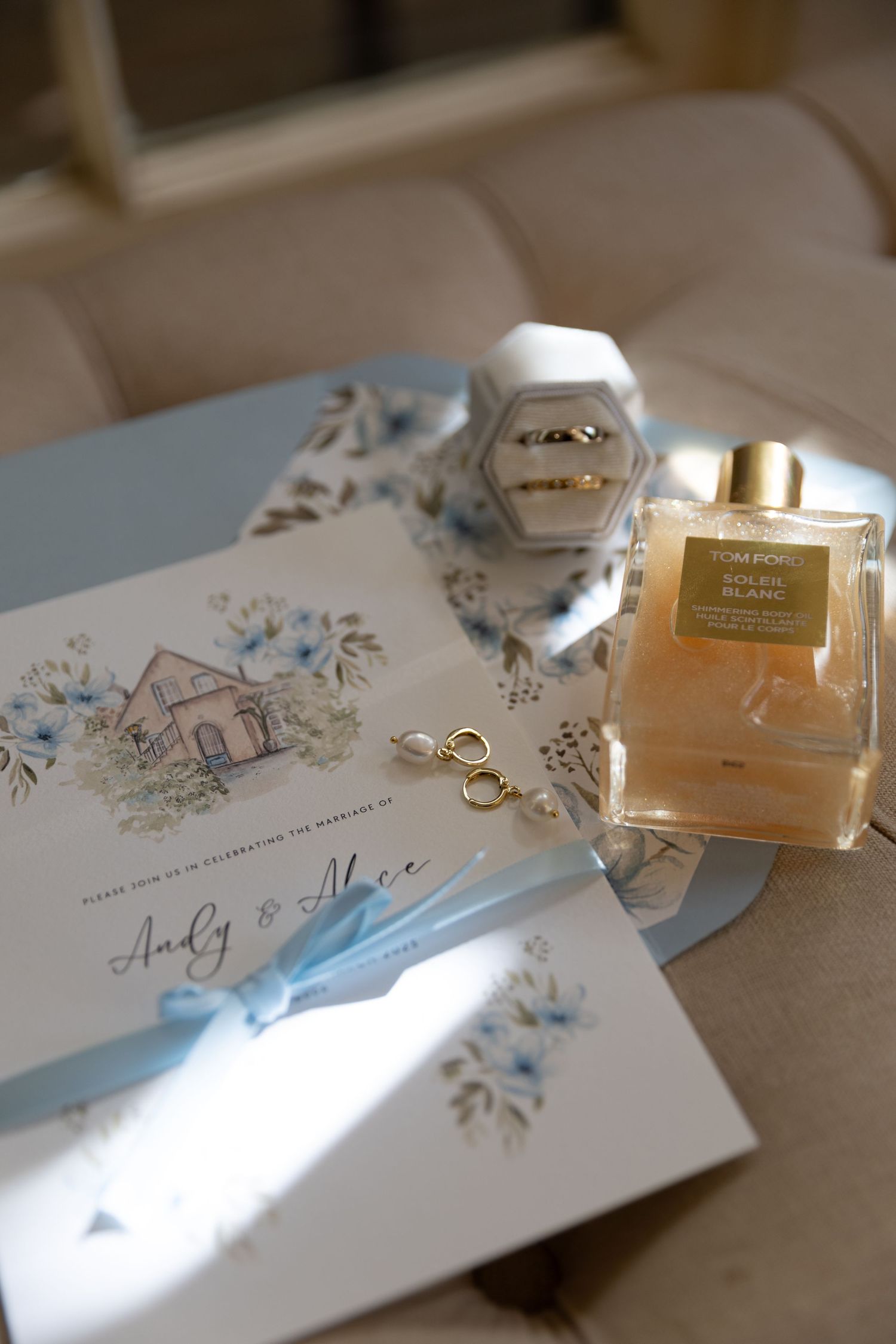 Wedding invitation with floral design alongside perfume bottle and ring box in soft lighting.
