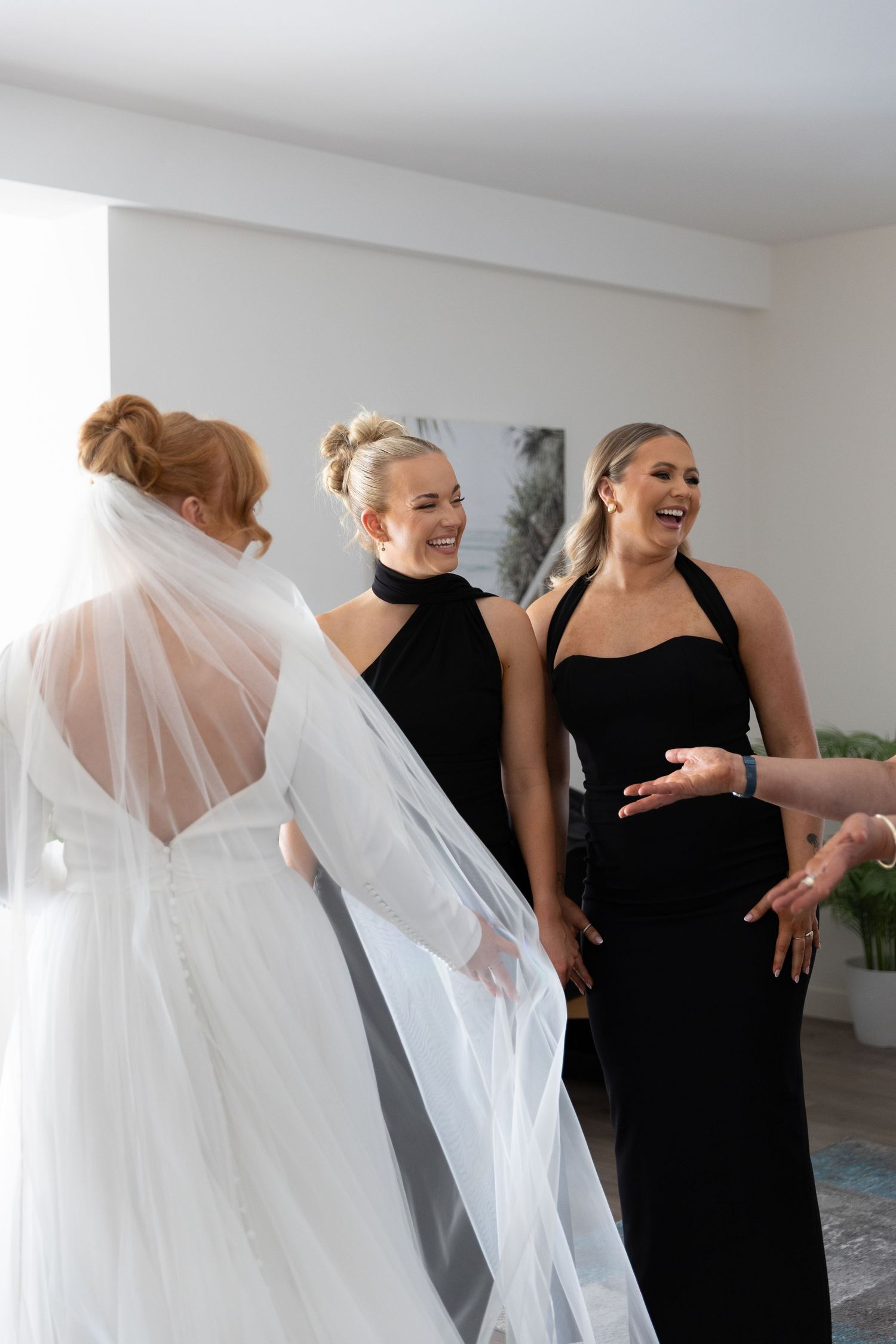 A bride in a flowing veil shares joyful moments with attendants wearing elegant black dresses in a bright indoor space.