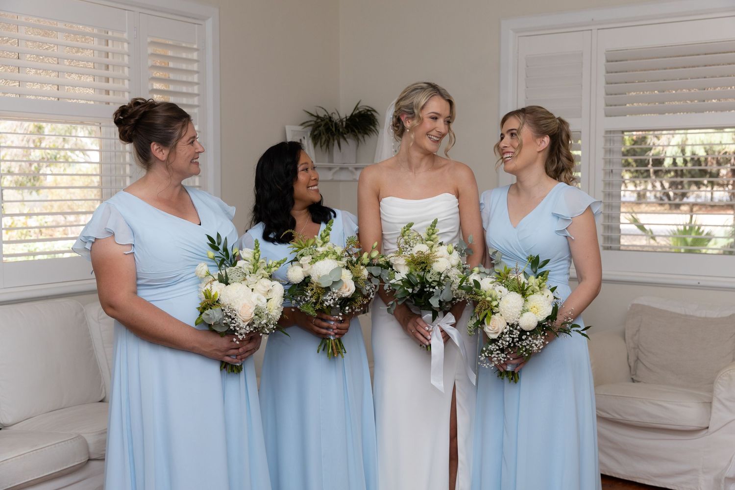 Bridal party in pale blue dresses holding white floral bouquets in a bright room.