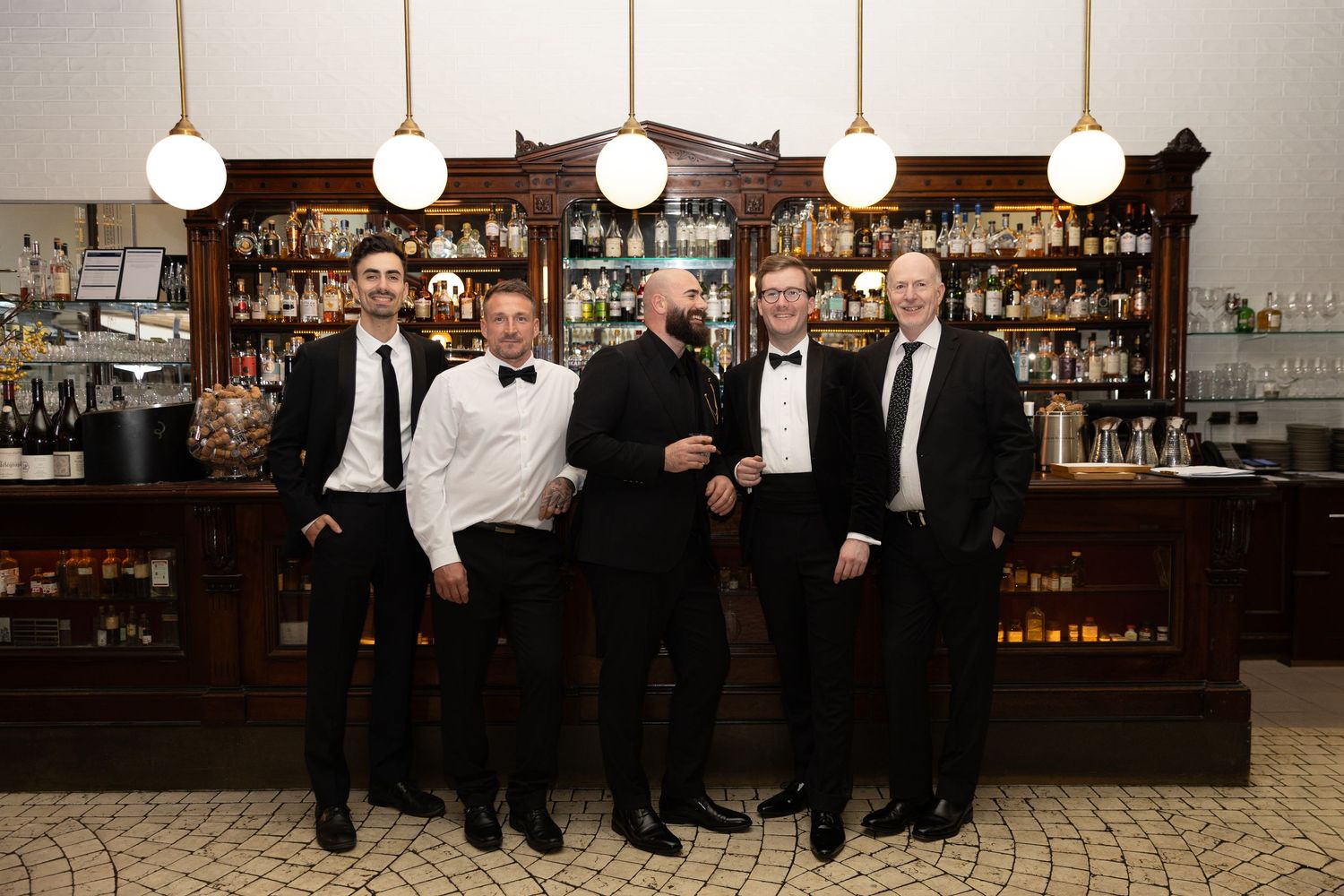 A group in formal black and white attire stands together at an elegant bar with vintage lighting and decor.