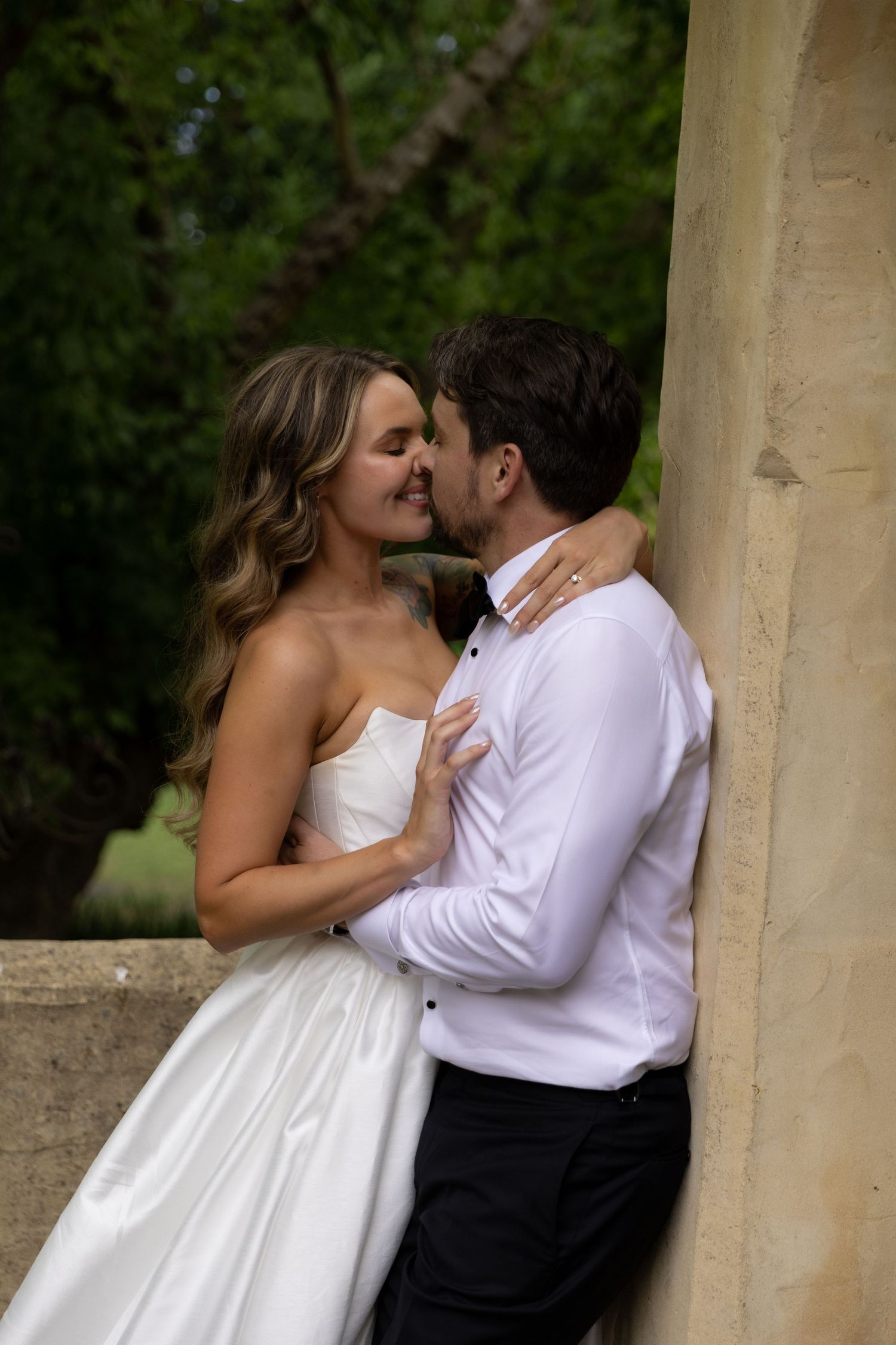 An intimate moment between a couple in wedding attire against a stone pillar outdoors.