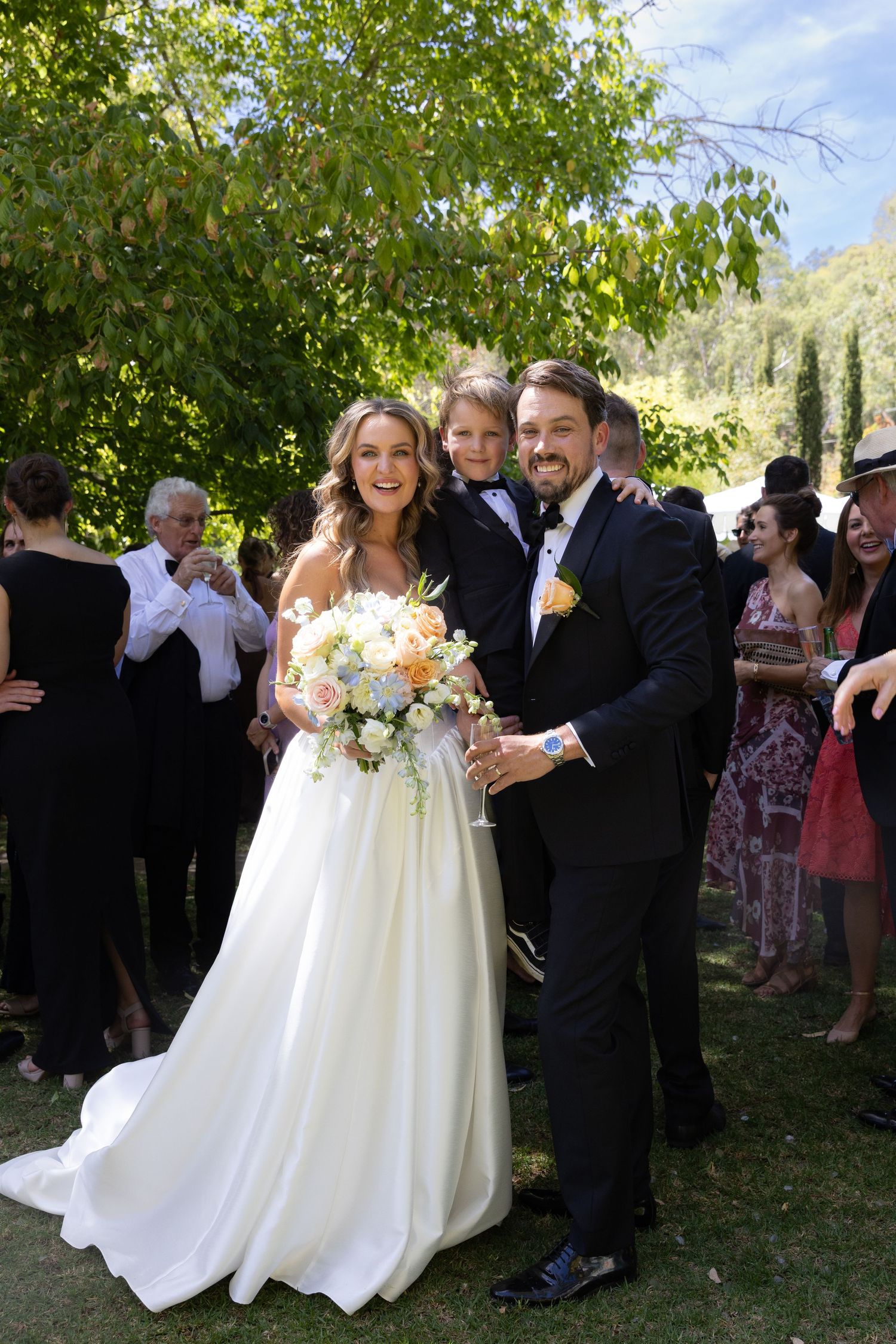 Outdoor wedding celebration with couple surrounded by guests under trees.