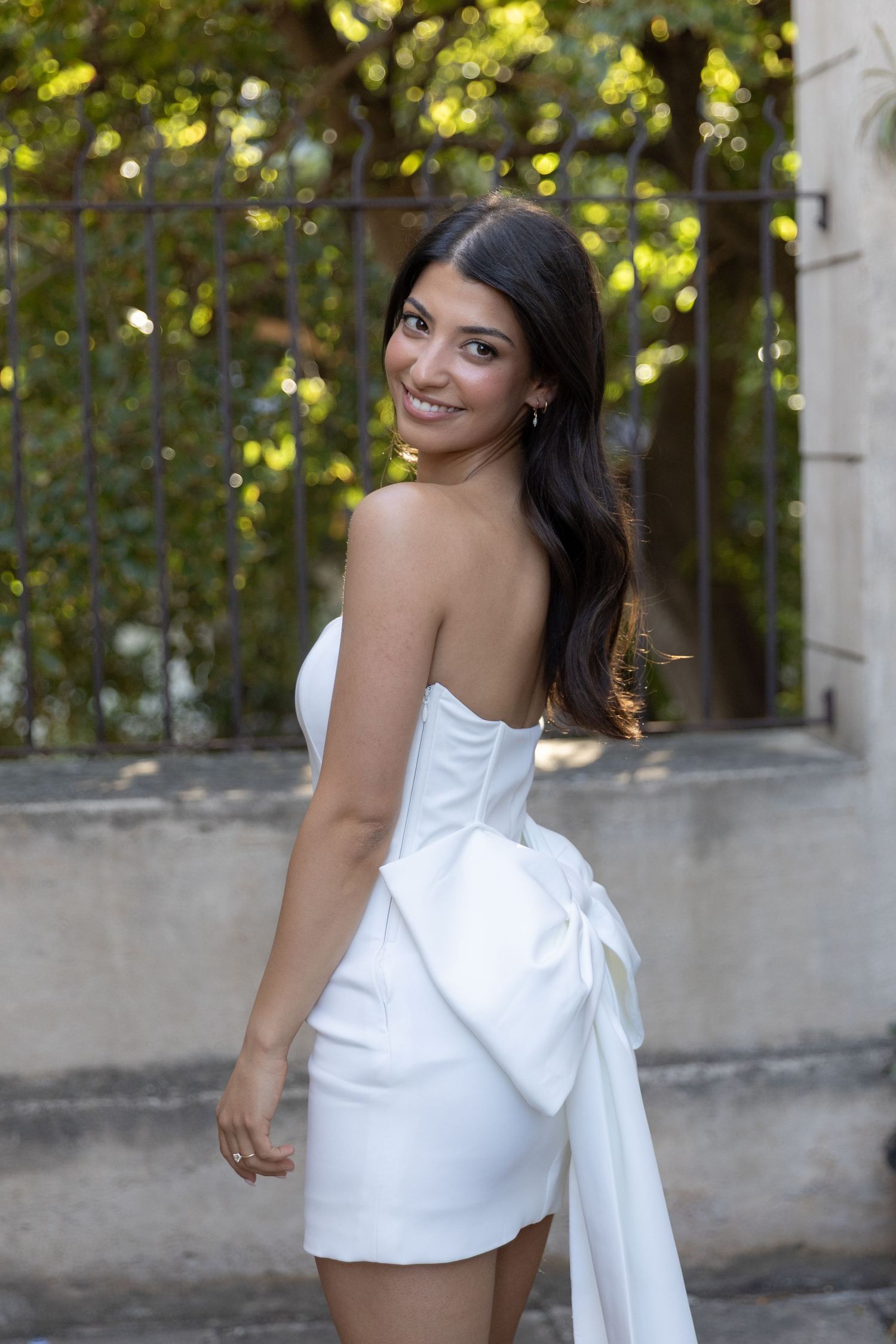 Woman in white strapless dress with bow detail smiles over shoulder in outdoor garden setting.