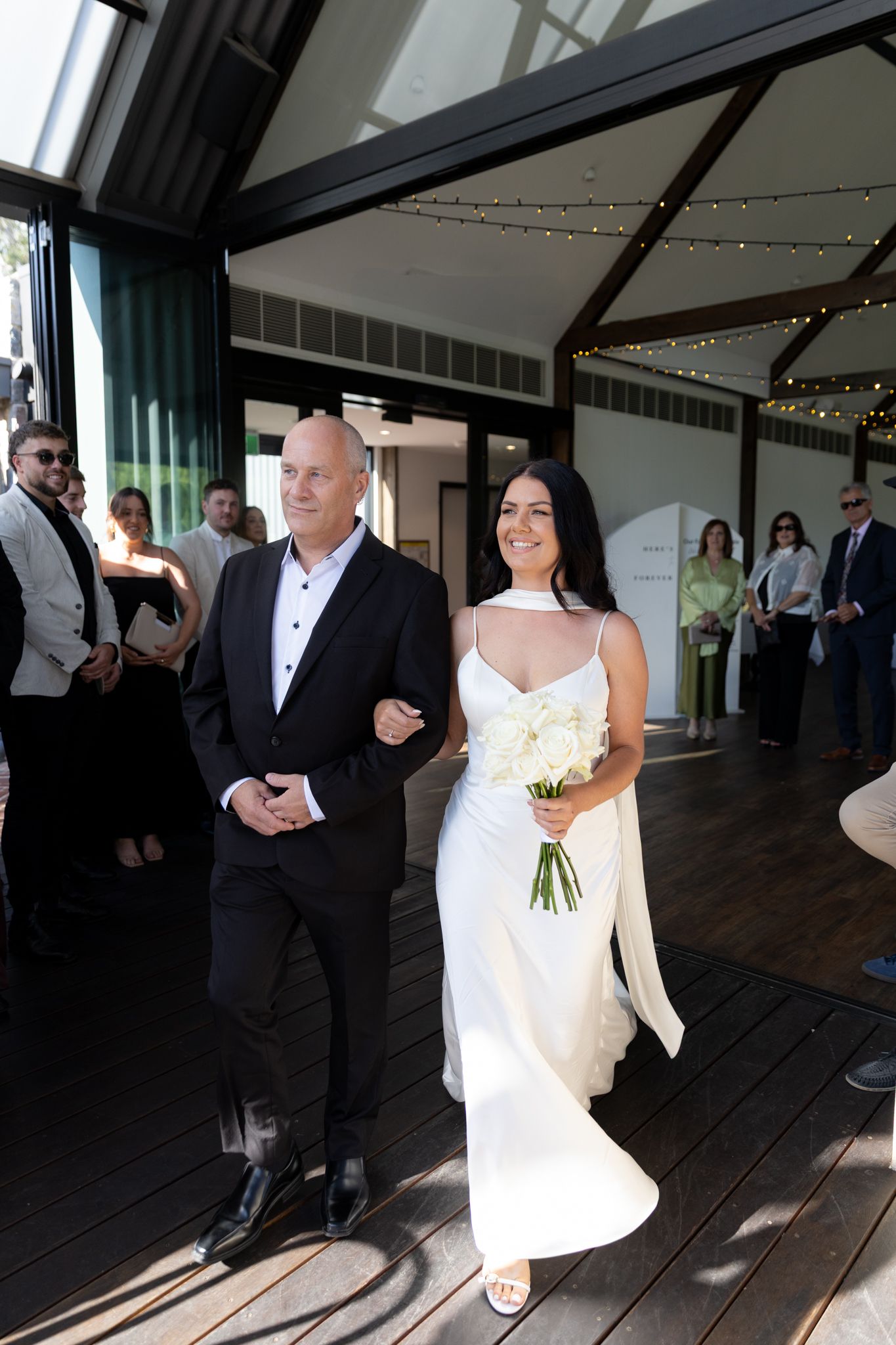 A bride in white gown walks alongside a man in formal wear under string lights at an indoor venue.