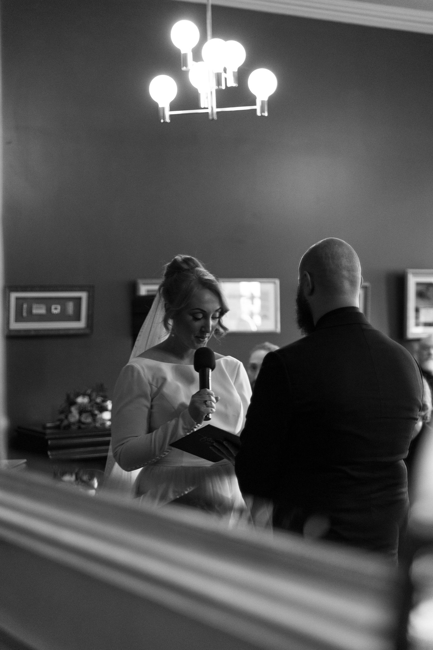 Black and white photograph of a wedding ceremony showing the couple sharing a moment under an elegant chandelier.
