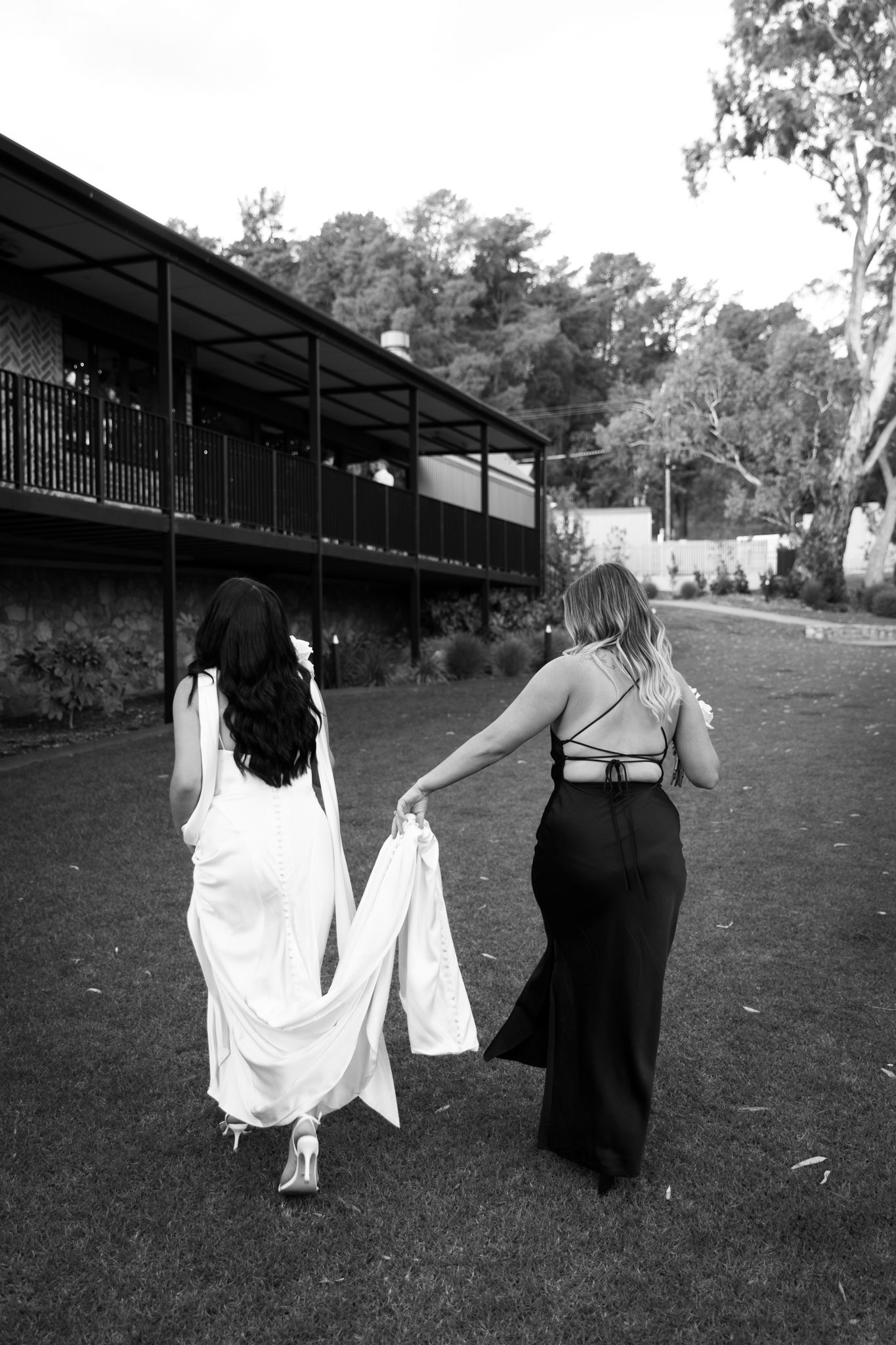 Two people walking on grass, one in white dress and one in black, with a building in background.