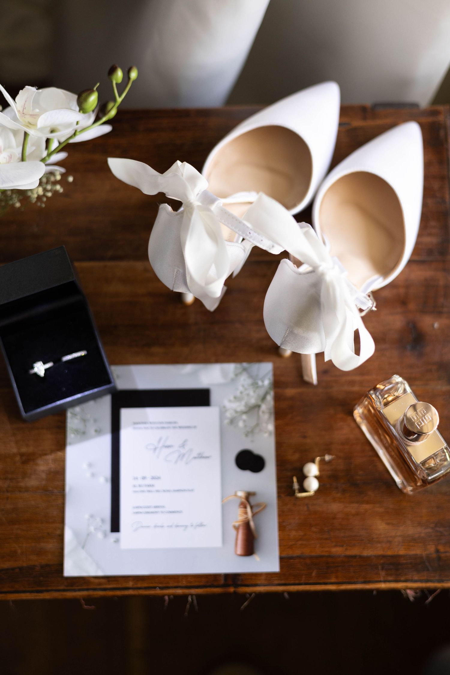 Wedding details flatlay with white heels, invitation, and floral accents on wooden surface.