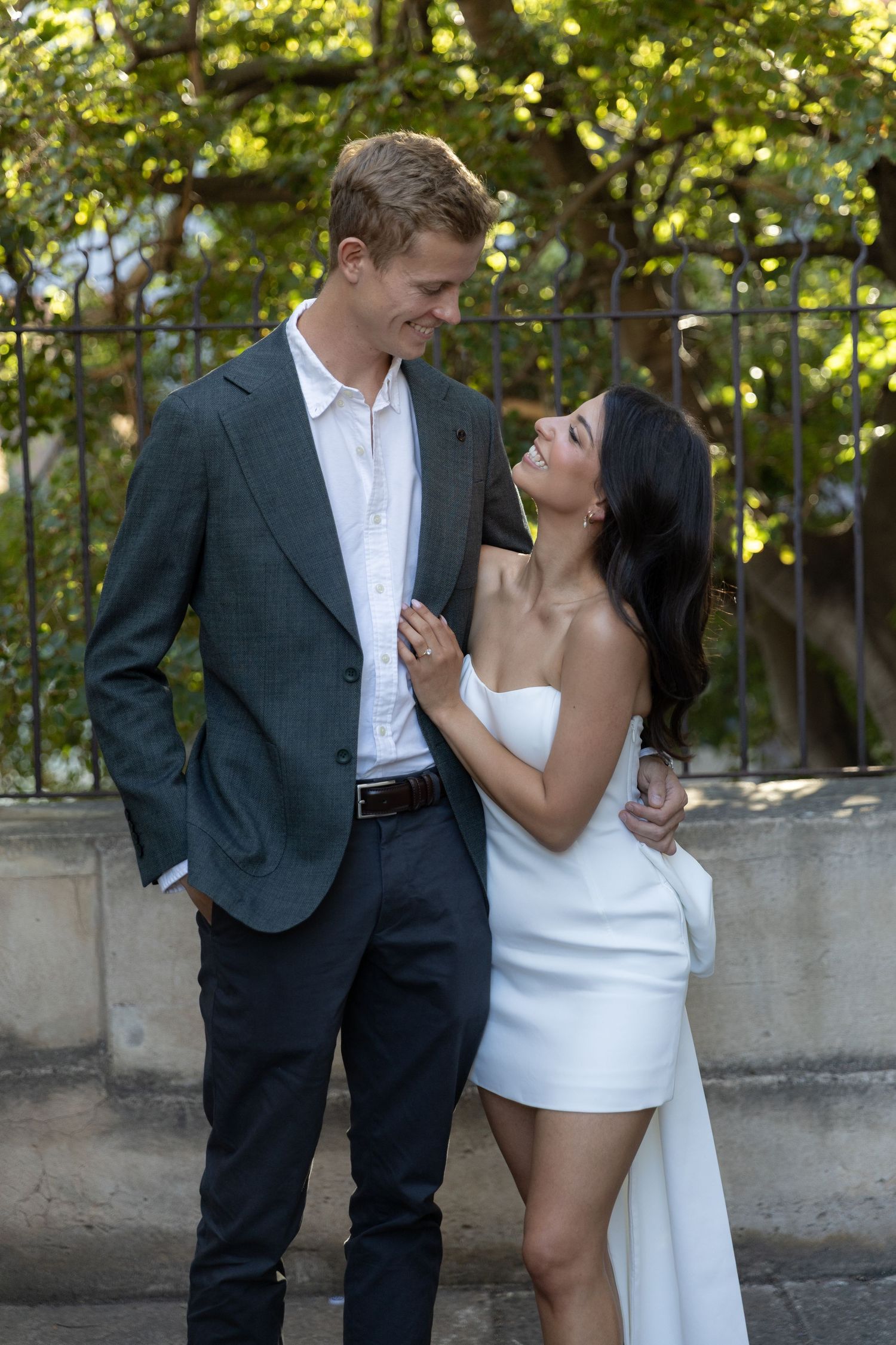 A couple shares a romantic moment outdoors in formal attire against a green garden backdrop.