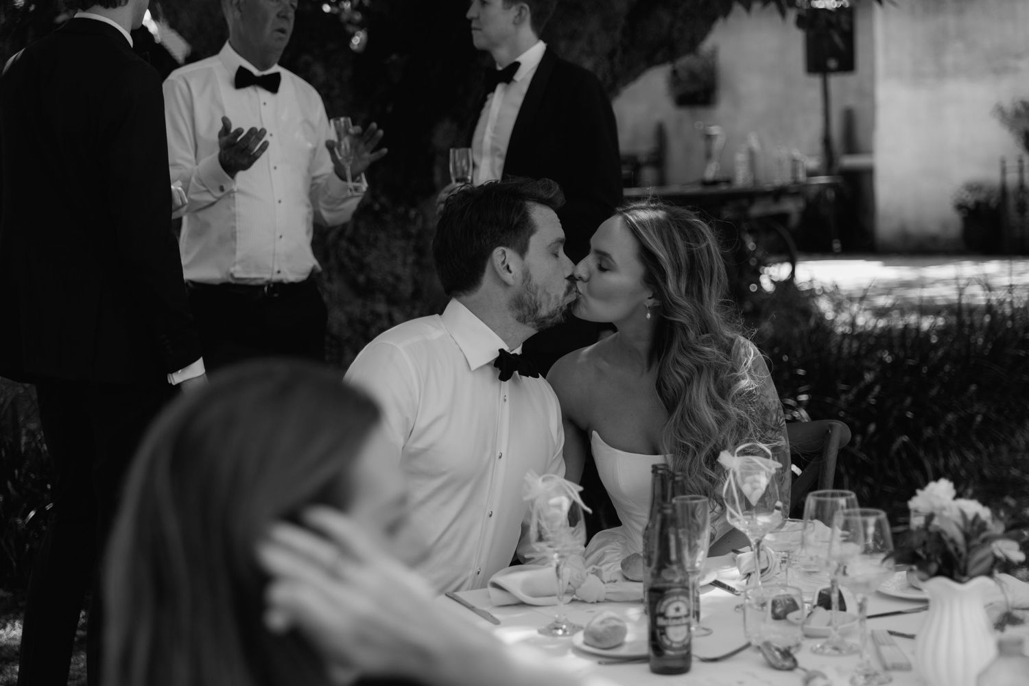 A candid black and white photo of wedding guests enjoying a reception dinner outdoors.
