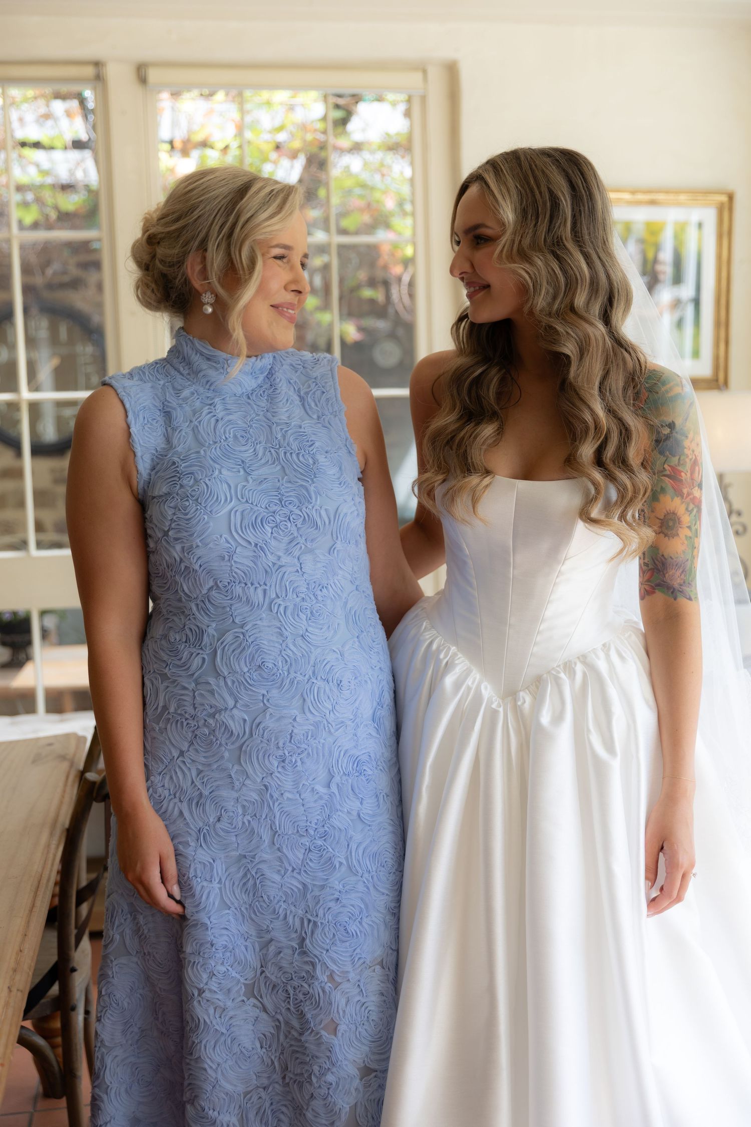 A bride in white poses with a companion wearing a blue lace dress in a sunlit room.