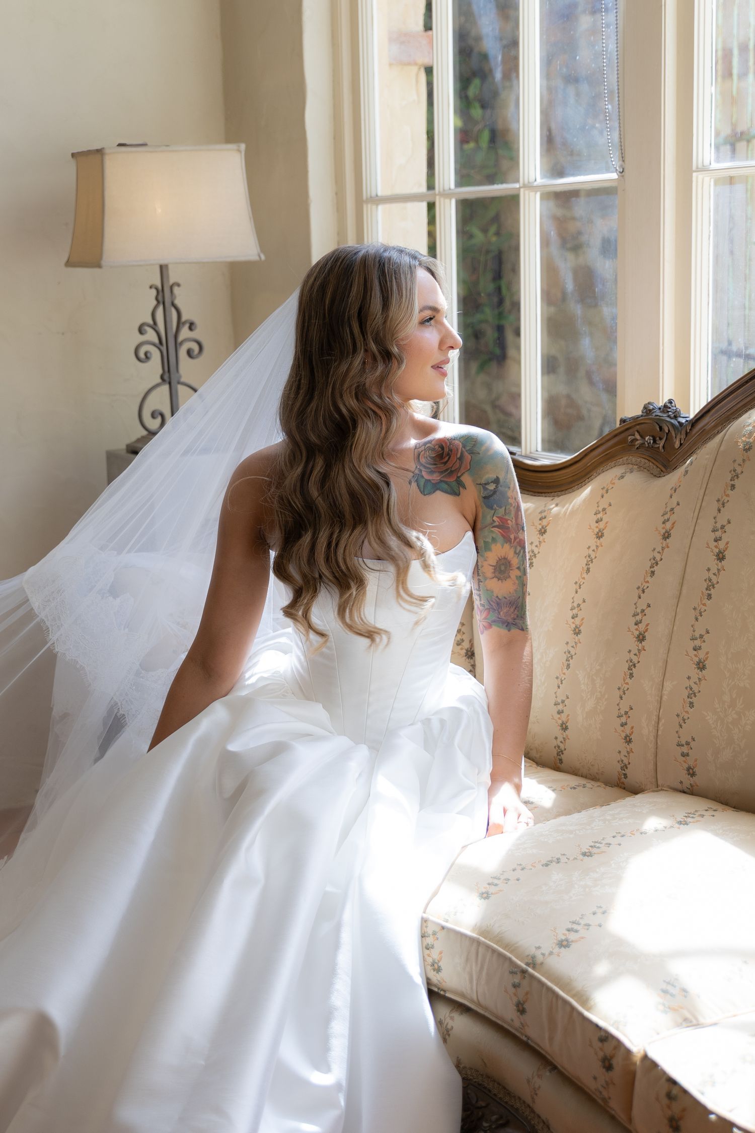 A bride in a strapless ballgown sits by a window, natural light illuminating her profile.