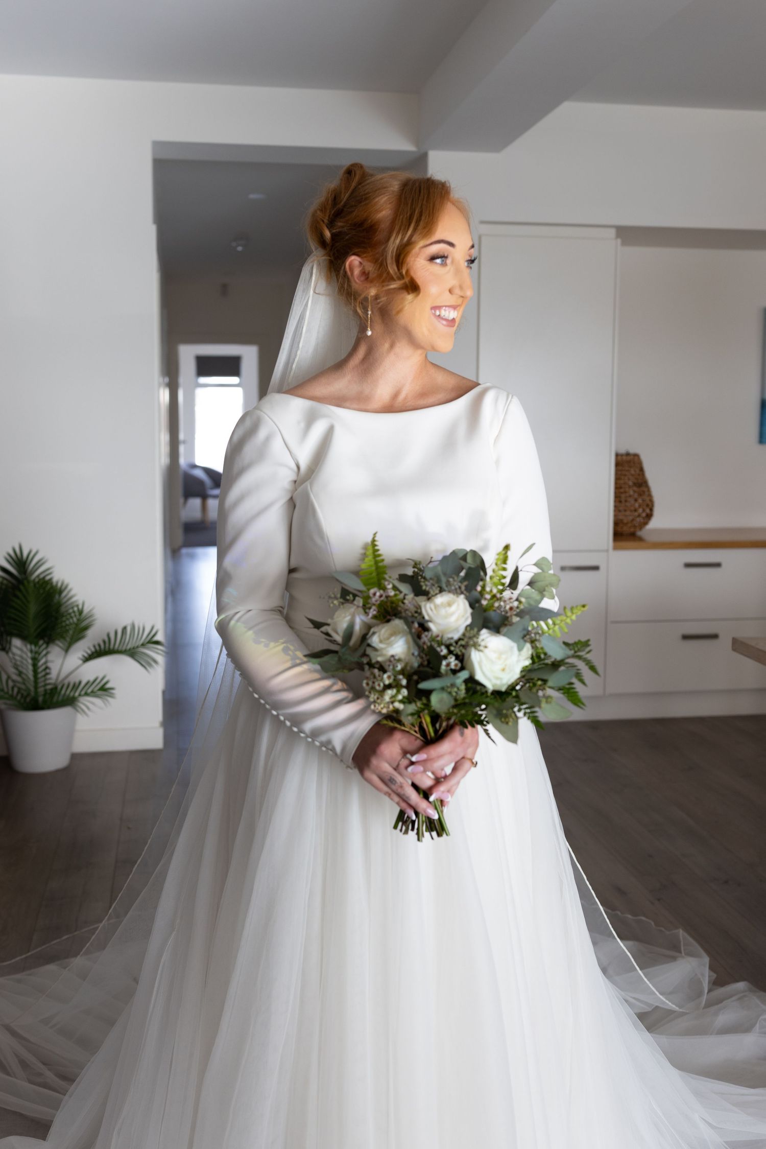 A bride in an elegant long-sleeved white gown and veil holds a romantic bouquet of white roses and greenery in a modern interior.