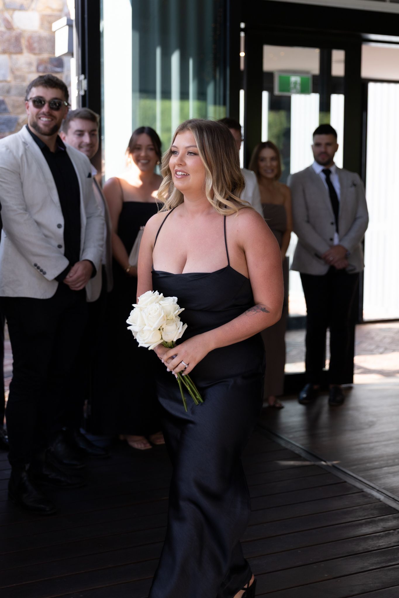 Bridesmaid in a black dress holding white flowers stands with wedding guests in a venue.