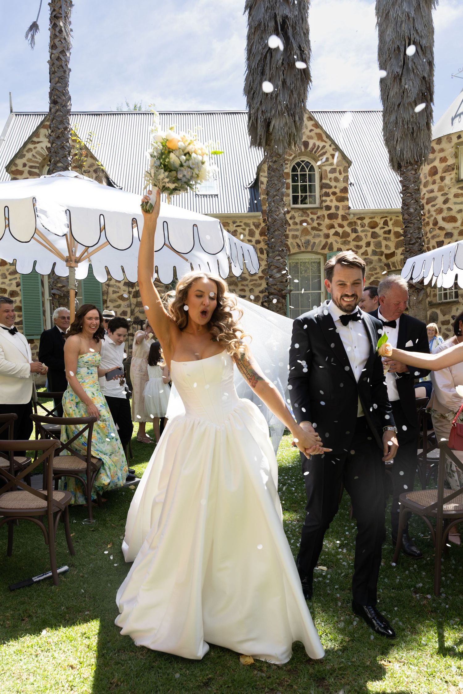 Newlyweds celebrate with joy outside a stone church as guests throw confetti in the sunshine.