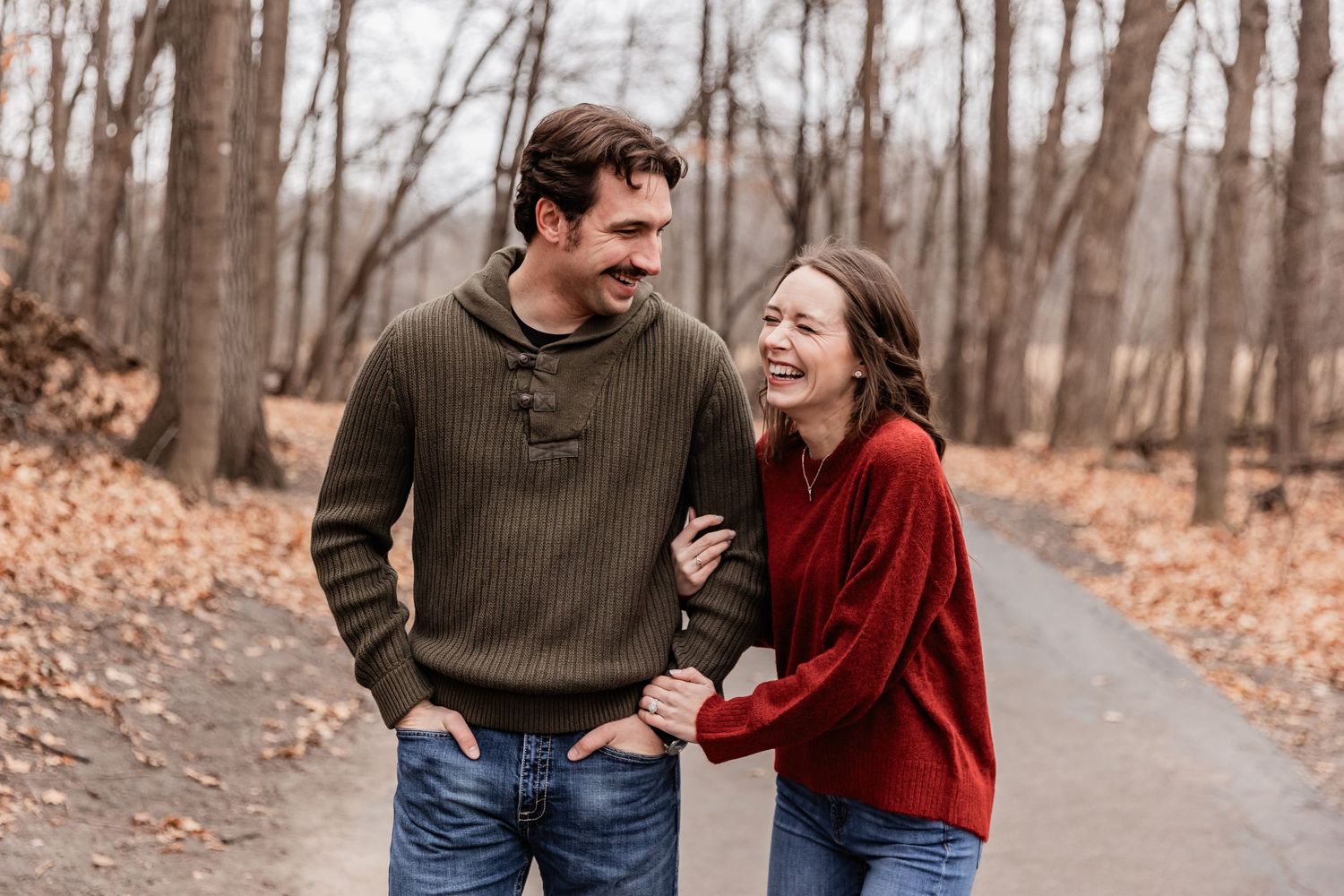 Young woman and he husband wear cozy sweaters as they laugh walking down a trail