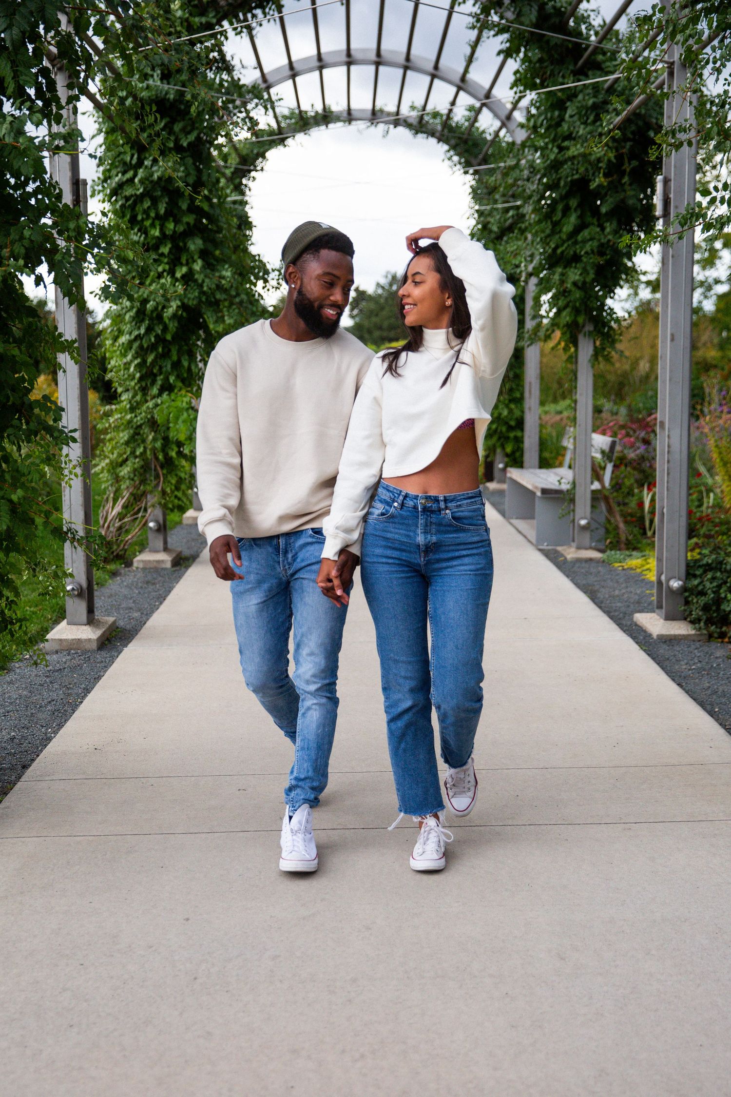 Ivan and his girlfriend walk holding hands under a white metal archway as the green bushes surround them