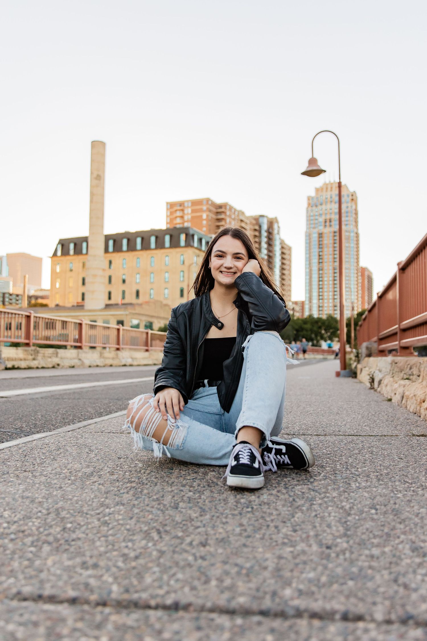 Sidney rests her head on her hand as she sits on Stone Arch Bridge with the city gleaming behind her