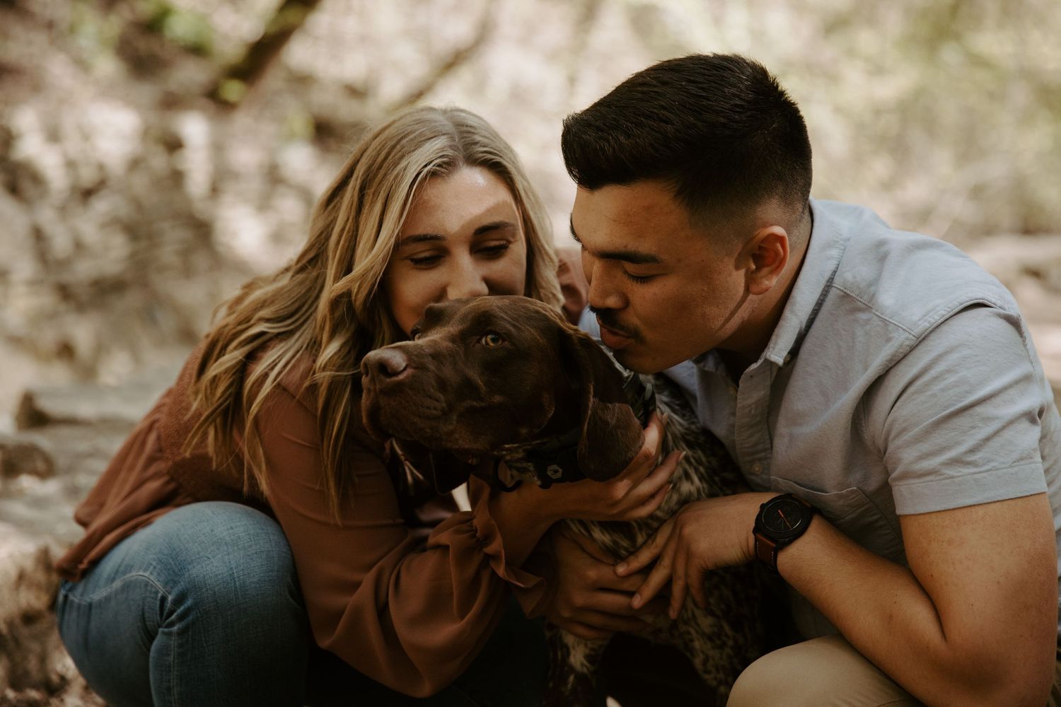Young engaged couple leans over to kiss their chocolate lab on the head