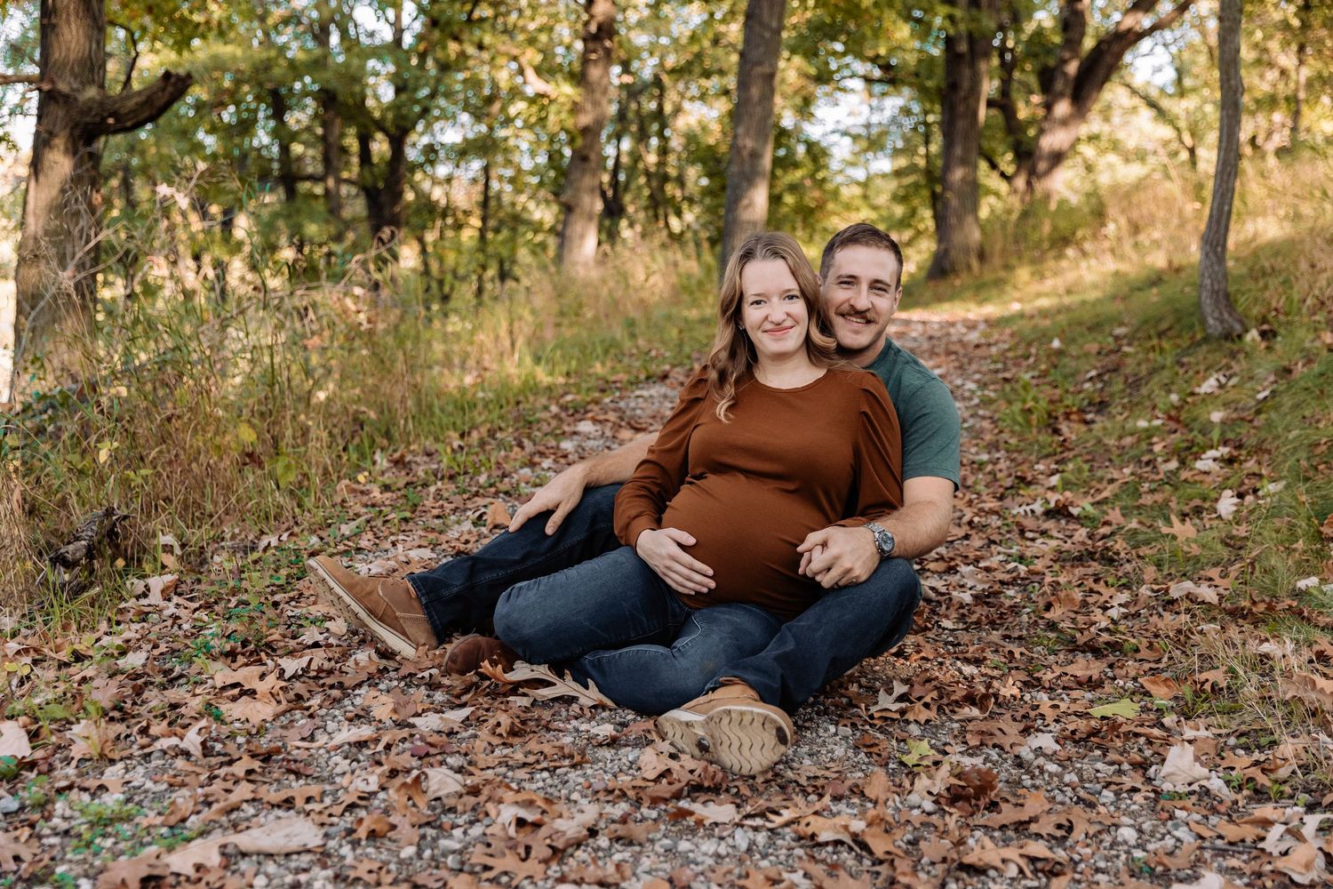 Pregnant woman sits on leaf covered ground and leans back into her husband with trees and tall grass surrounding them