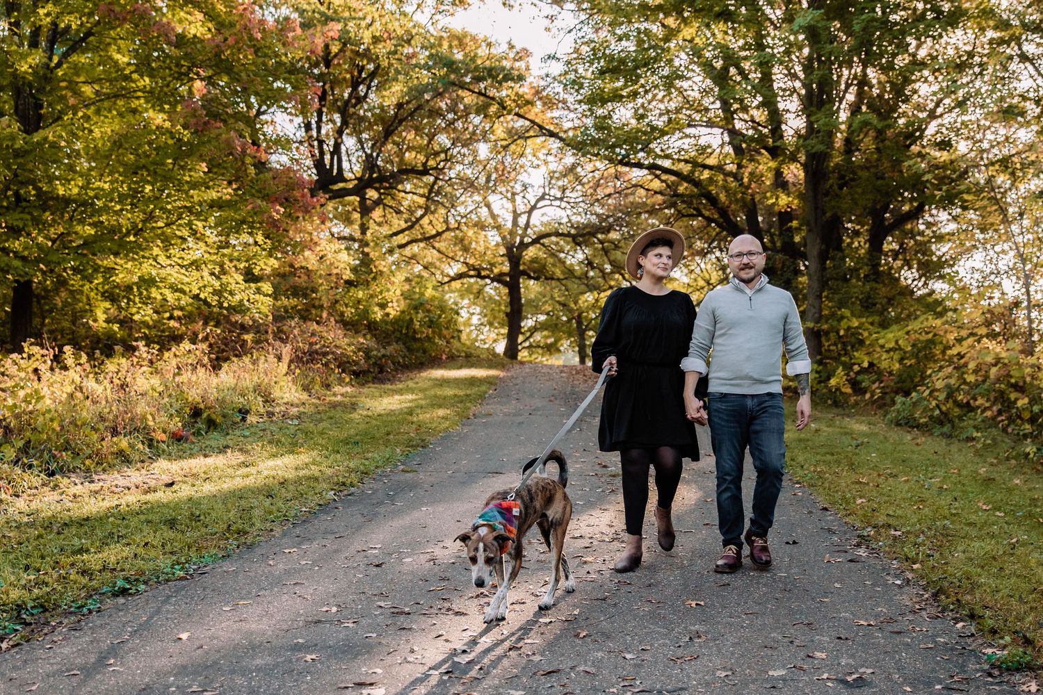 Husband and wife walk their boxer puppy down a paved trail with the sunset beaming through the trees