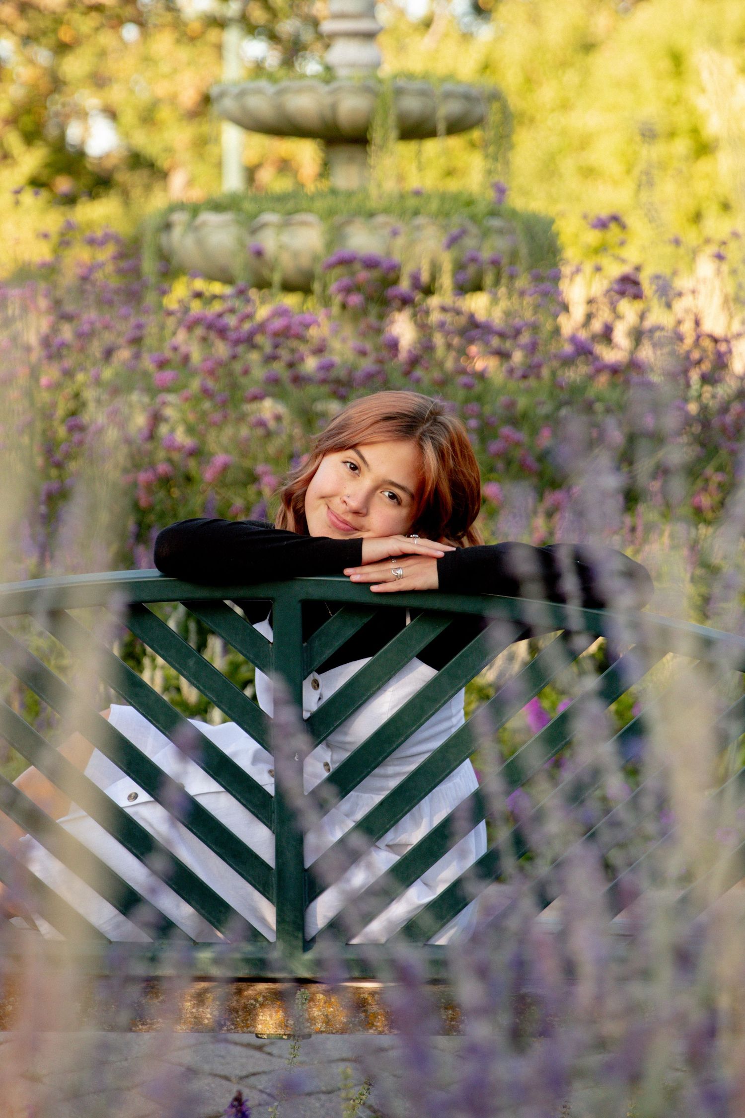 Confident seniors rests her head on her hands as she looks back over a bench surrounded by flowers and a water fountain