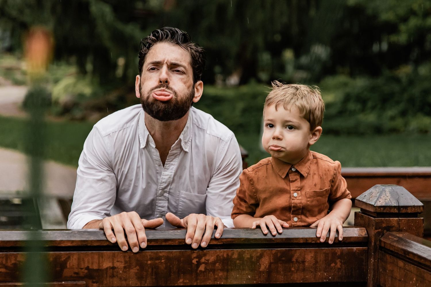 Dad and son make silly faces as they kneel on a bridge together